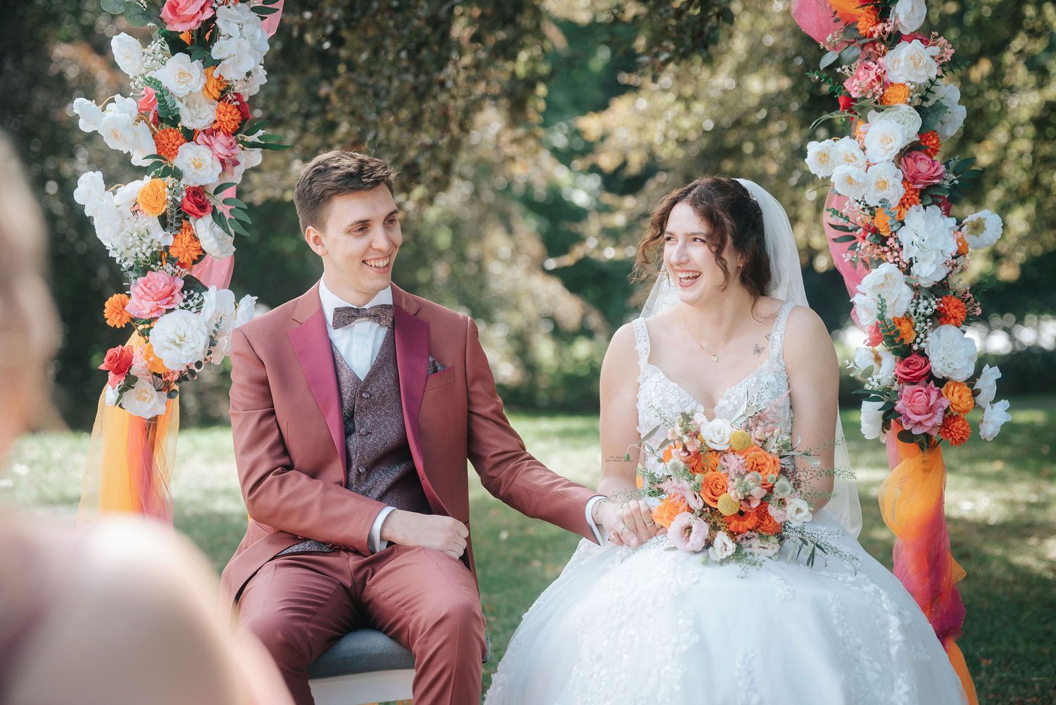 Smiling bride in a white lace gown and groom in a burgundy suit hold hands during an outdoor wedding ceremony with colorful floral arrangeme