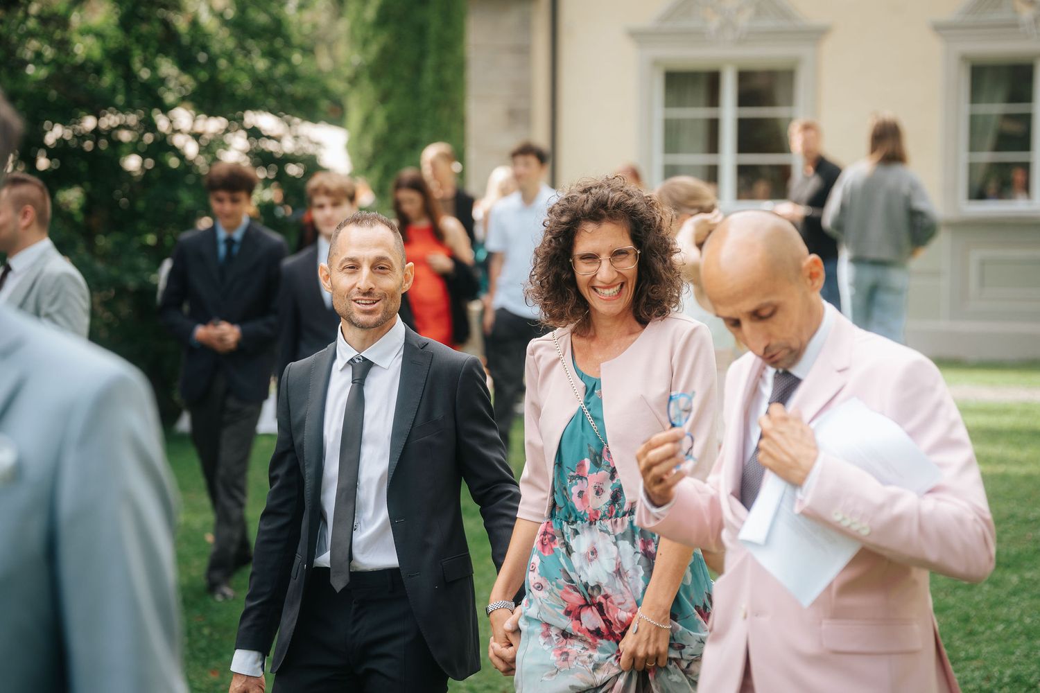Smiling wedding guests in elegant attire mingle on a sunny lawn, with a man in a dark suit and a woman in a floral dress.