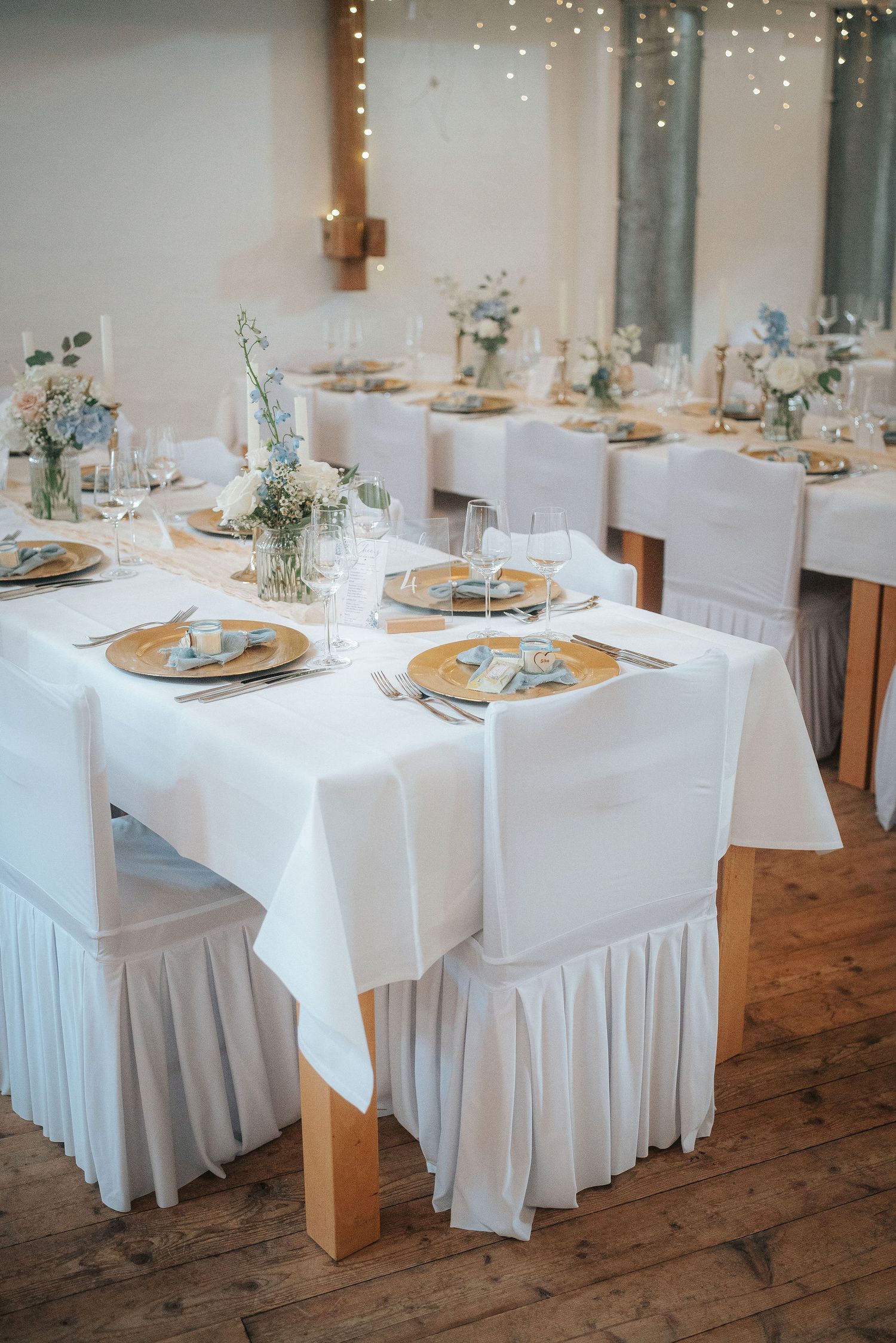 Rustic wedding reception tables with white linens, gold chargers, blue and white floral centerpieces, and chair covers on a wood floor.
