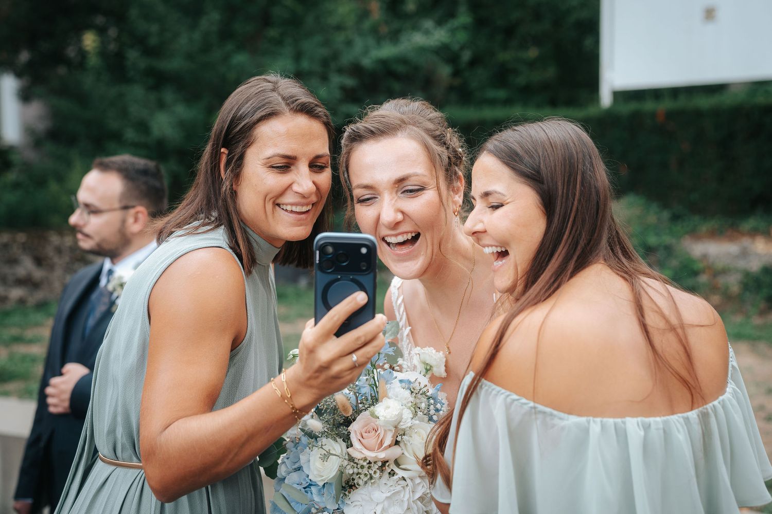 Three smiling women in sage green dresses gather close to look at a smartphone at a wedding, one holding a pastel bouquet.