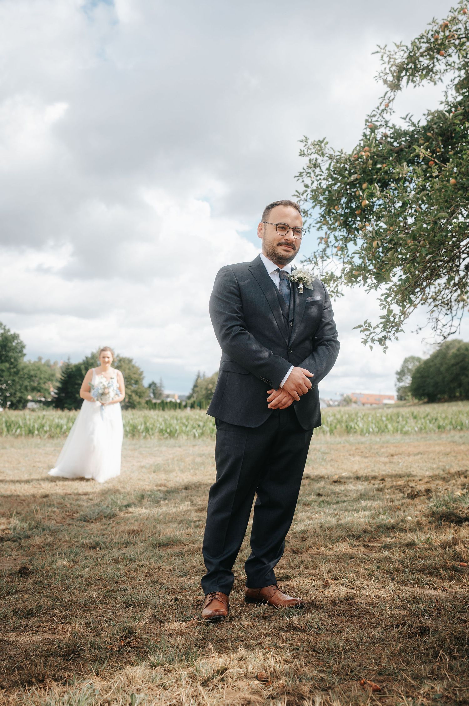 Groom in a navy suit and glasses waits in a grassy field for a first look while the bride in a white gown approaches behind him.