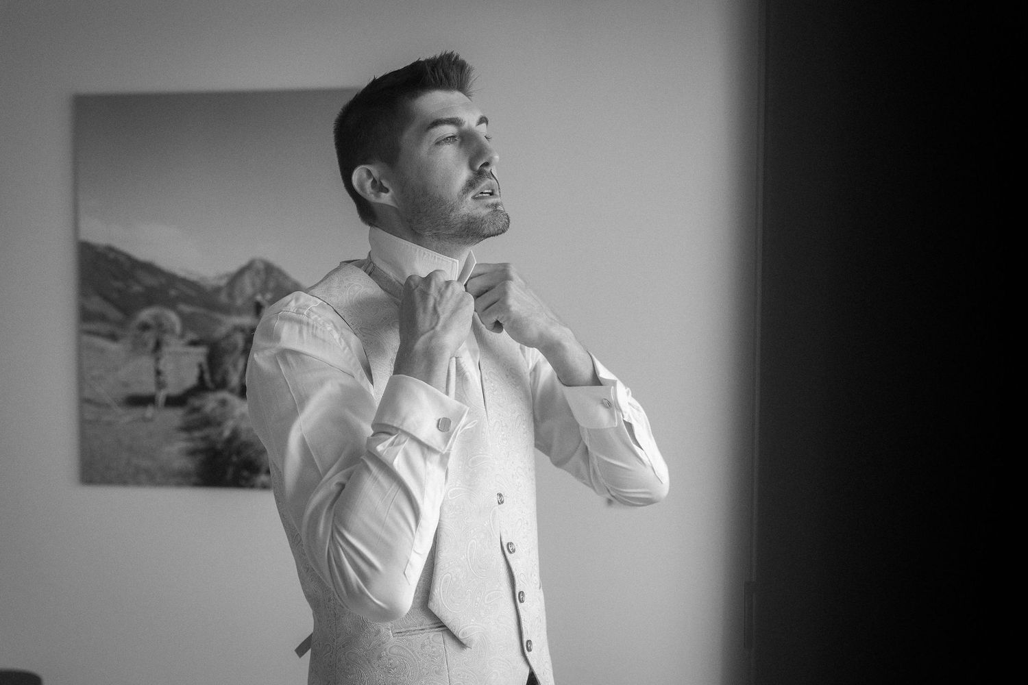 Groom in a white dress shirt and vest adjusts his collar by a window while getting ready for his wedding in a black and white photo.