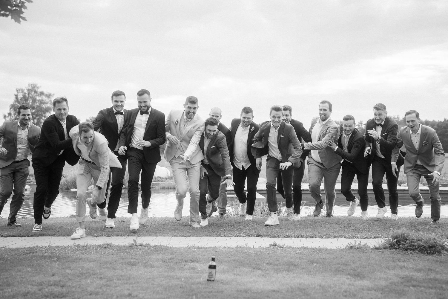 Black and white photo of a group of groomsmen in suits running playfully toward the camera across a grassy lawn with trees behind them.