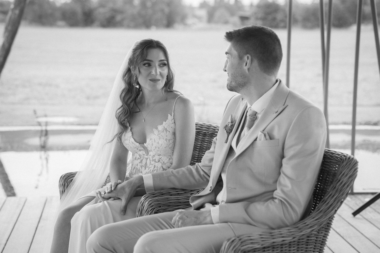 Black and white photo of a bride in a lace gown and veil sitting beside a groom in a suit on wicker chairs at an outdoor wedding ceremony.
