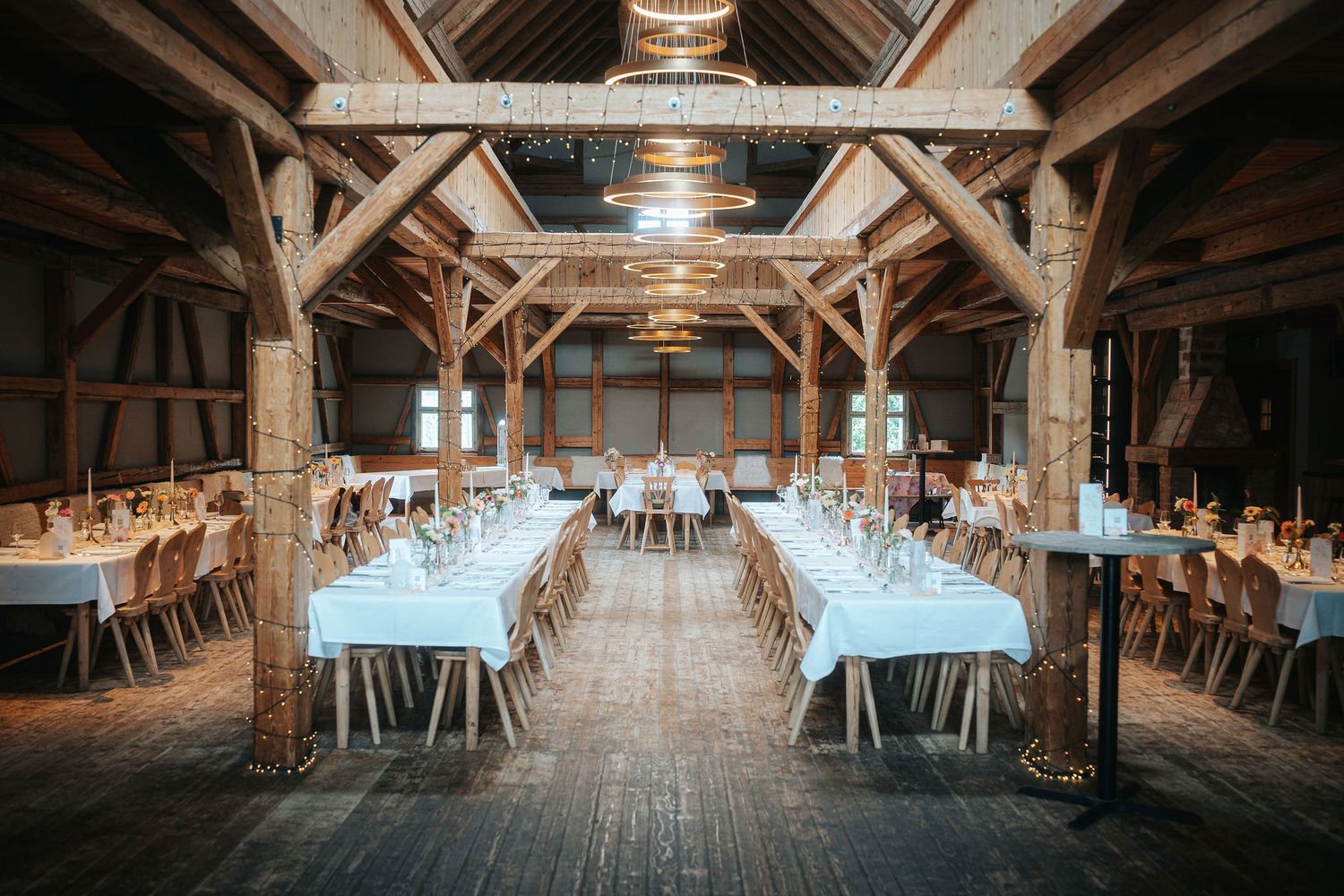 Rustic barn wedding venue with long tables set with white linens, wooden chairs, exposed timber beams and chandeliers.