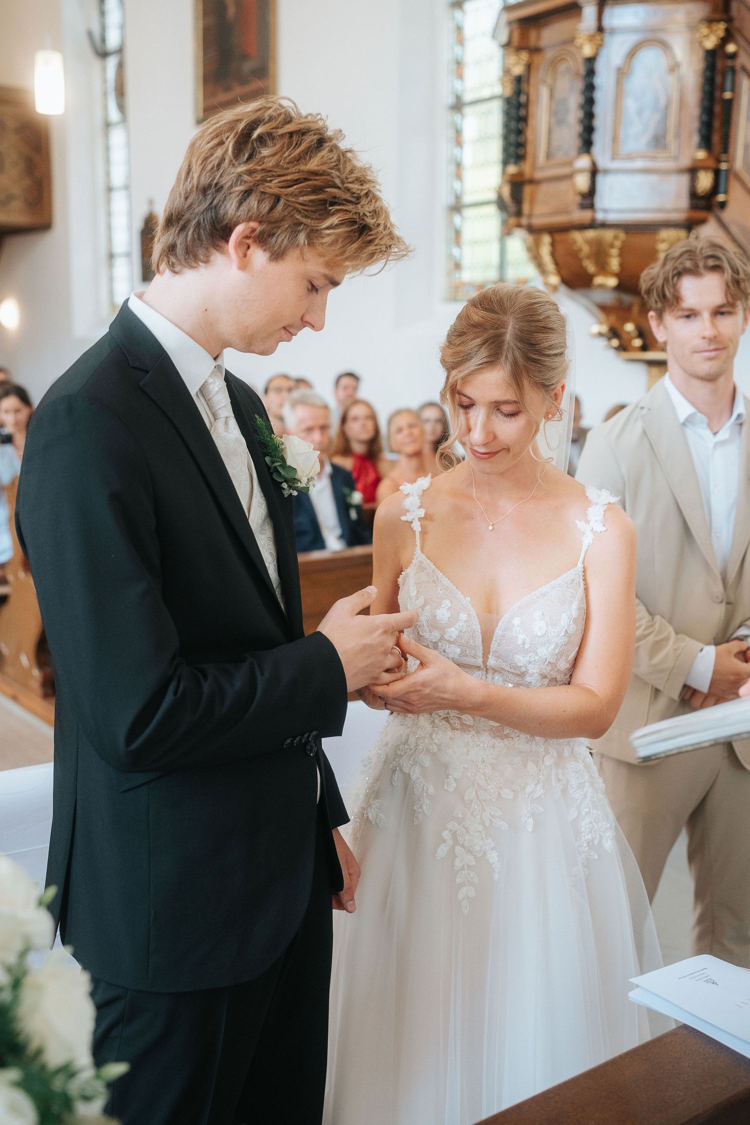Bride in a lace wedding gown and groom in a black suit exchange vows during a church ceremony as guests look on.