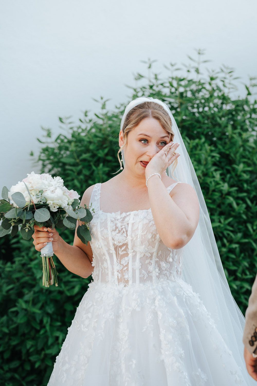 Emotional wedding photography series showing bride in white lace dress wiping tears while holding bouquet against greenery.