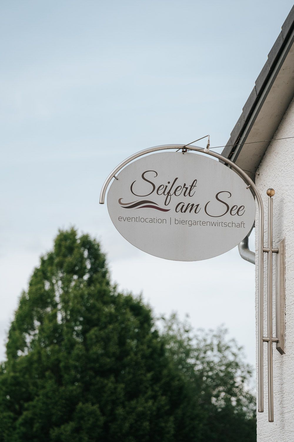 A storefront sign in German reading 'Leben am See' with elegant lettering mounted on a pole against trees.