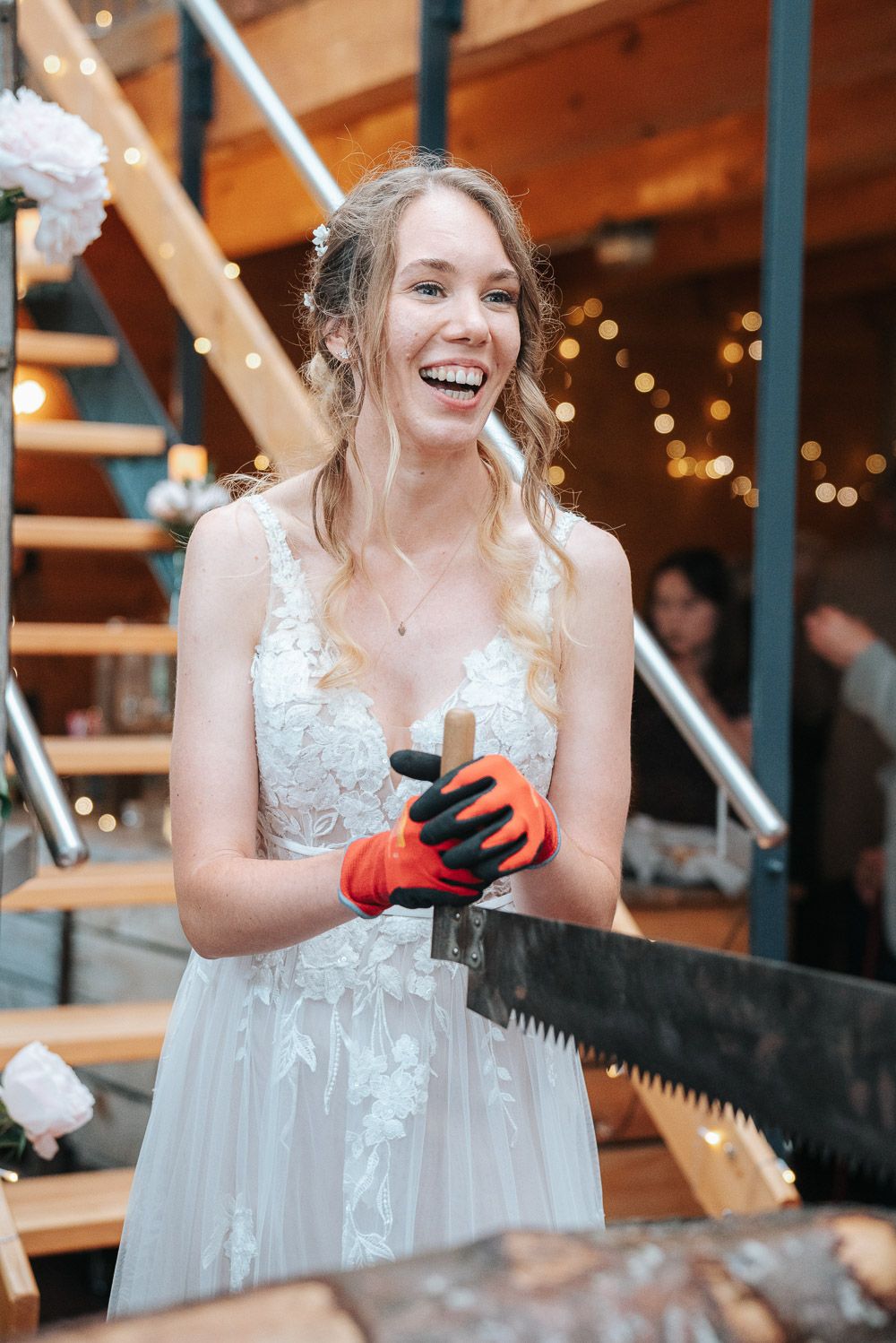 A joyful bride in a white lace dress plays games at a rustic indoor venue with warm lighting and wooden beams.