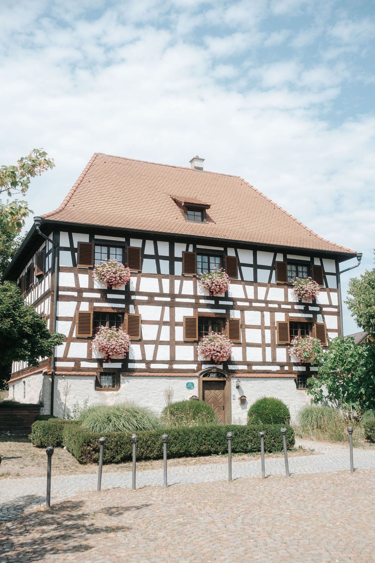 Half-timbered house with a red tile roof and flower boxes on the shuttered windows, framed by a trimmed hedge on a sunny cobblestone square.