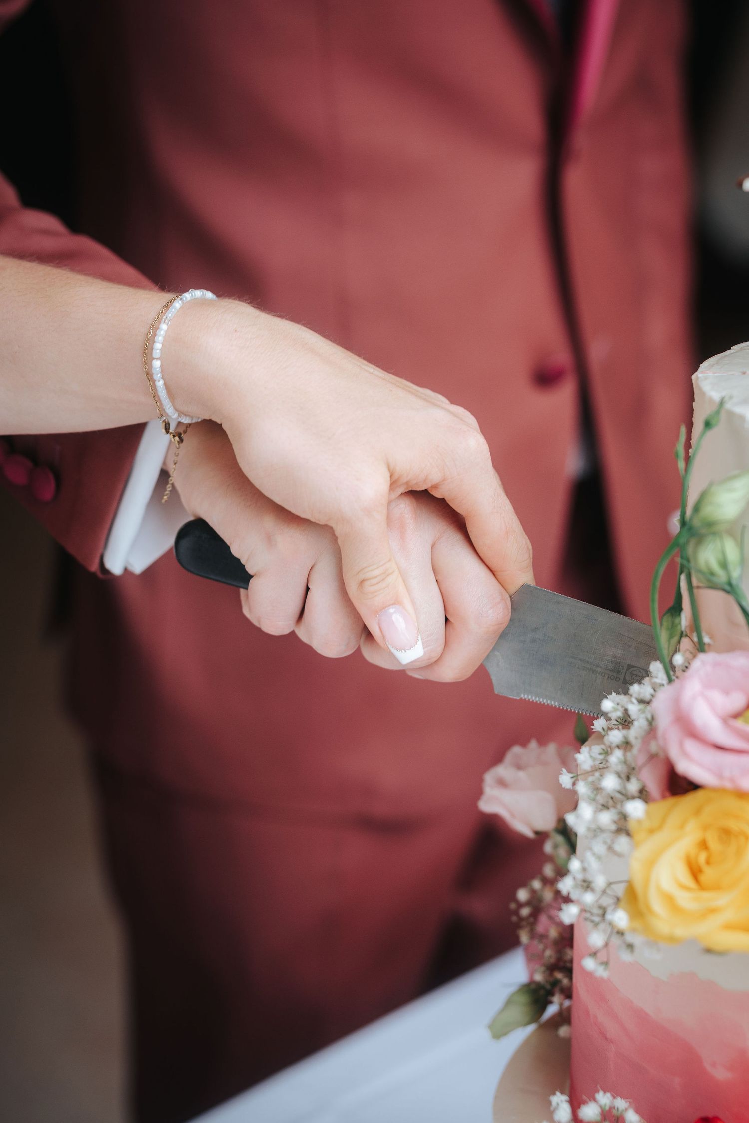Two hands holding a knife together to cut a pink wedding cake decorated with yellow roses, pink flowers, and baby's breath.