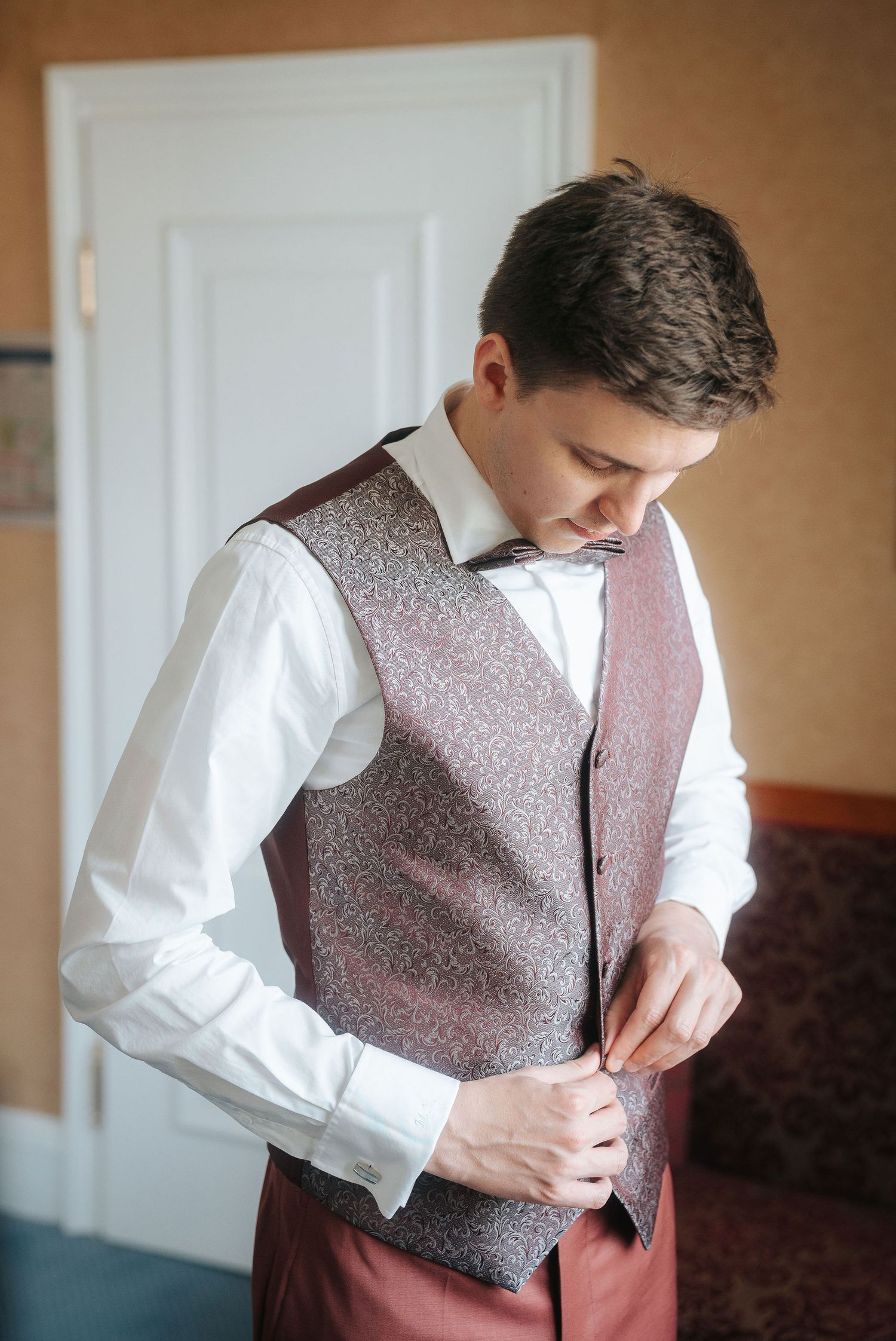 Groom in a white shirt and patterned burgundy vest buttons his waistcoat while getting ready for his wedding indoors.