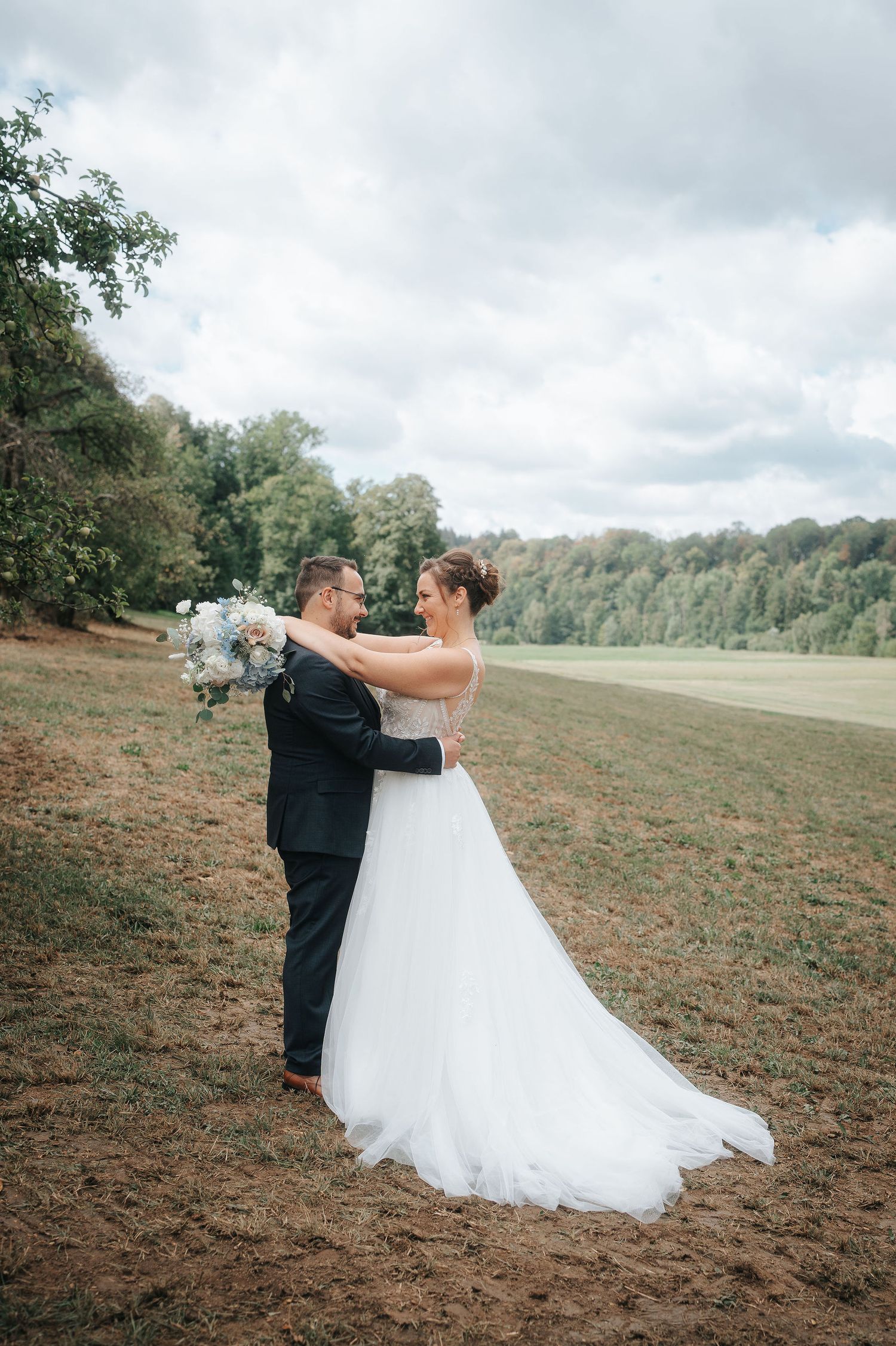 Bride in a white tulle gown and groom in a dark suit embrace on a grassy hillside holding a bouquet, with trees and cloudy sky behind.