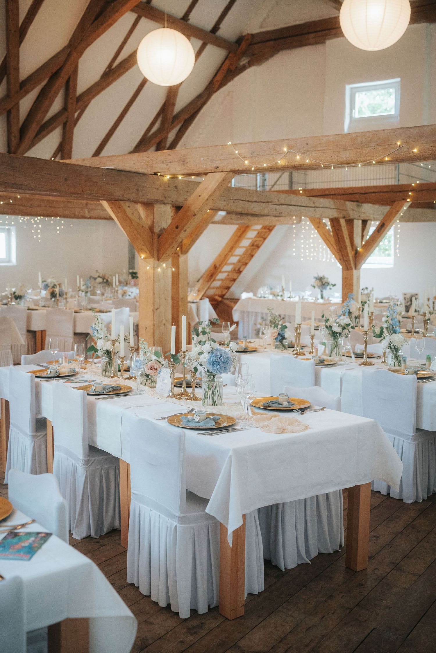 Rustic barn wedding reception with white draped tables, blue floral centerpieces, string lights and exposed wooden beams.