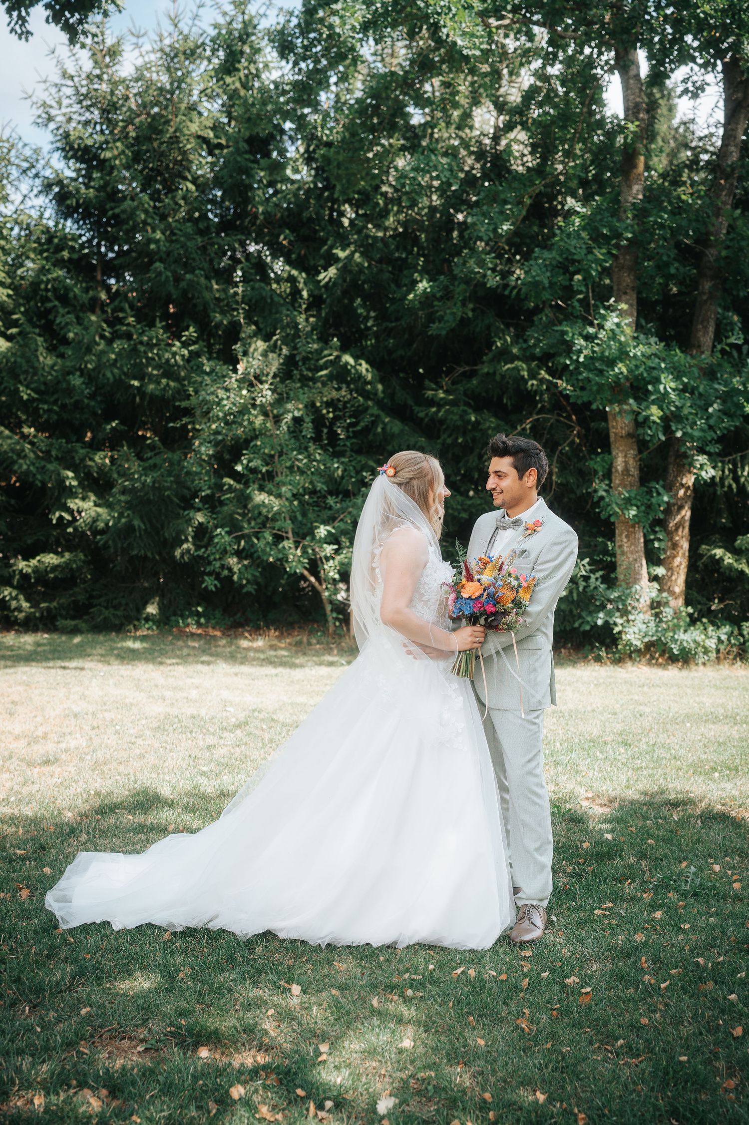 A bride in a white gown and veil embraces a groom in a light gray suit holding a colorful bouquet on a sunlit lawn framed by trees.