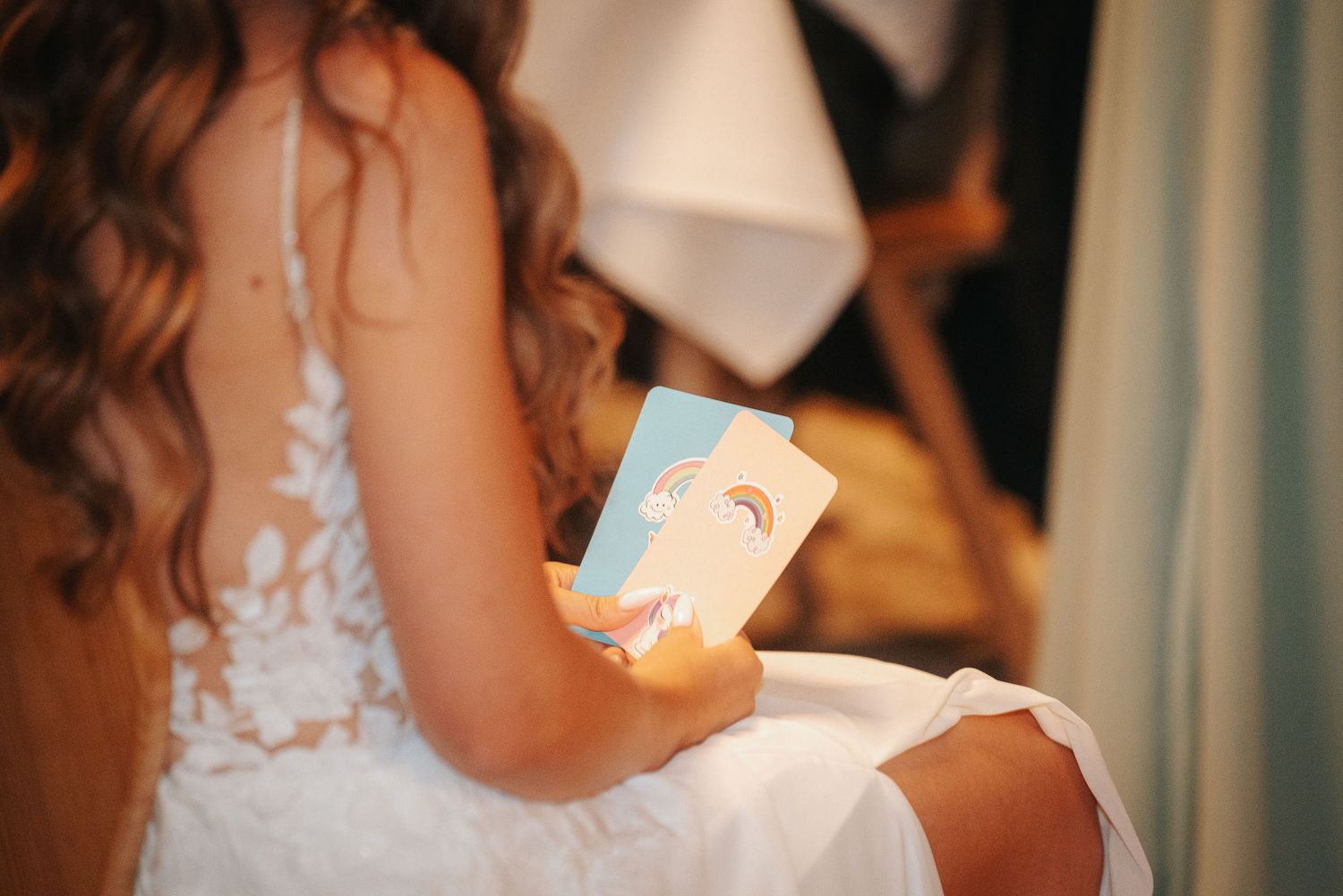 Bride in a white lace wedding dress with long wavy hair, seen from behind, holding a small card with a rainbow illustration.
