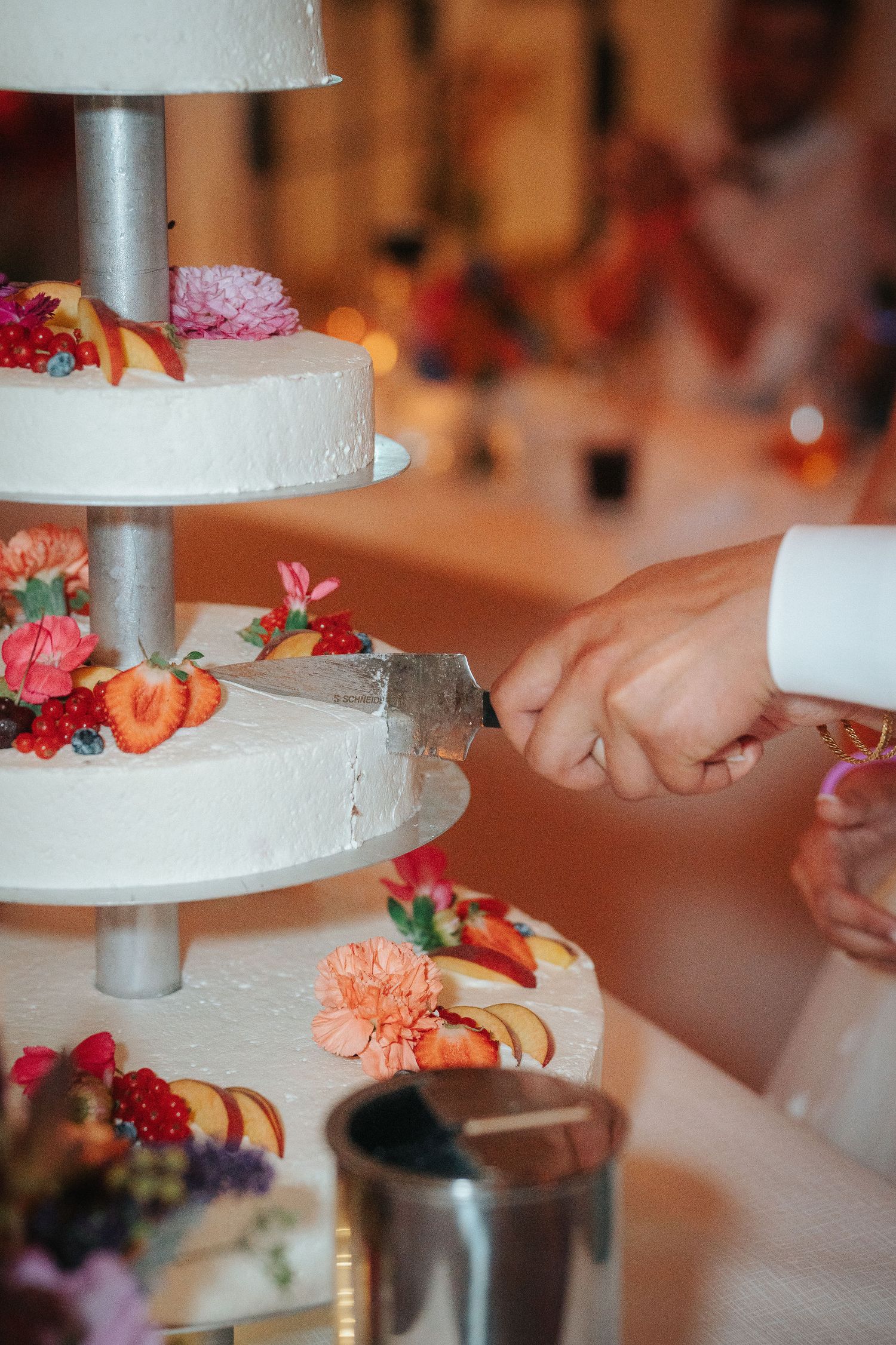 Couple cutting a tiered white wedding cake decorated with fresh flowers, peach slices, and berries during a reception.