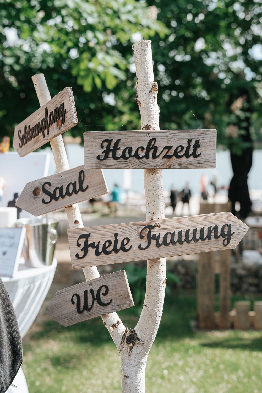 Wooden directional signs with German text are displayed on natural birch branches at an outdoor wedding decoration setup.