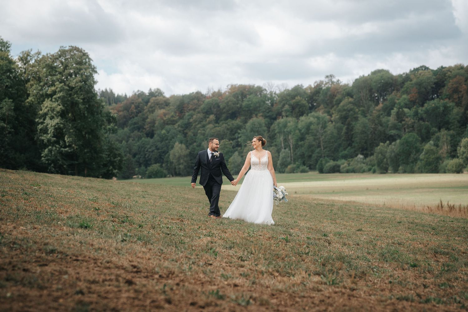 Bride in a white gown and groom in a dark suit hold hands while walking across a grassy meadow with wooded hills in the background.