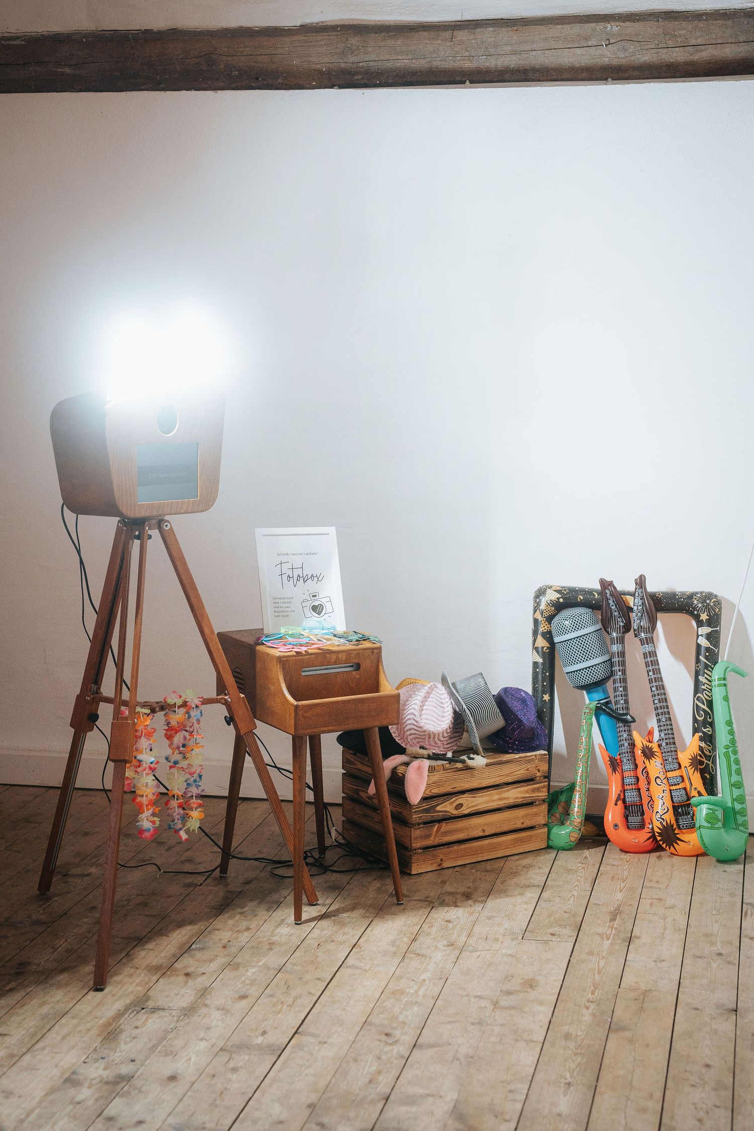 Vintage wooden tripod photo booth with bright flash beside a crate of hats and inflatable guitar props on a rustic wooden floor.