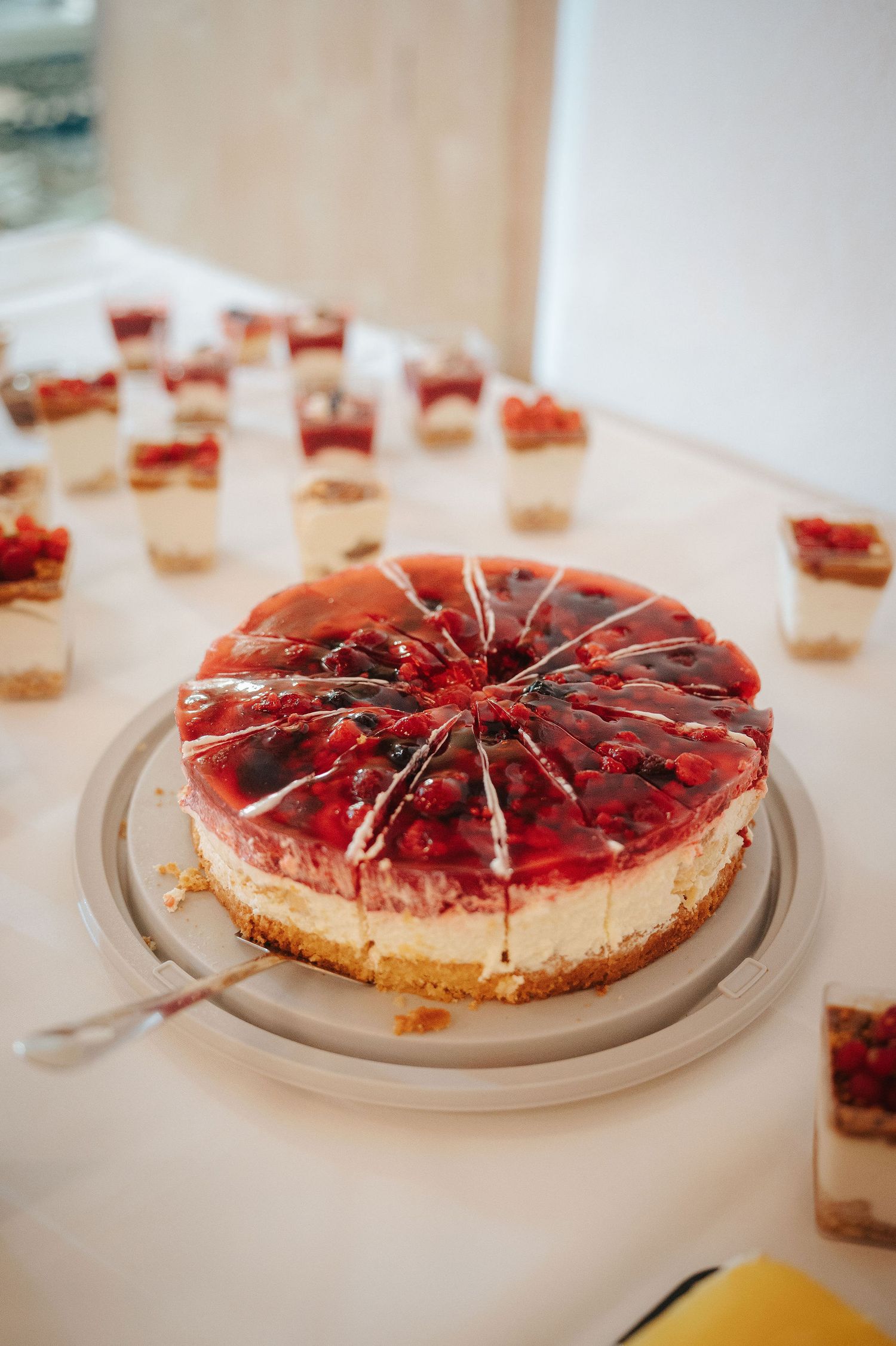 Sliced berry cheesecake with glossy red fruit glaze on a serving plate, surrounded by individual dessert cups on a buffet table.