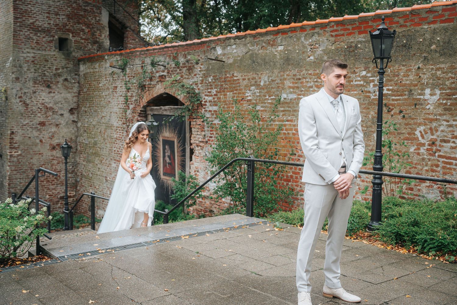 Groom in a light gray suit waits on a wet stone terrace as the bride in a white gown approaches from behind during a first look.