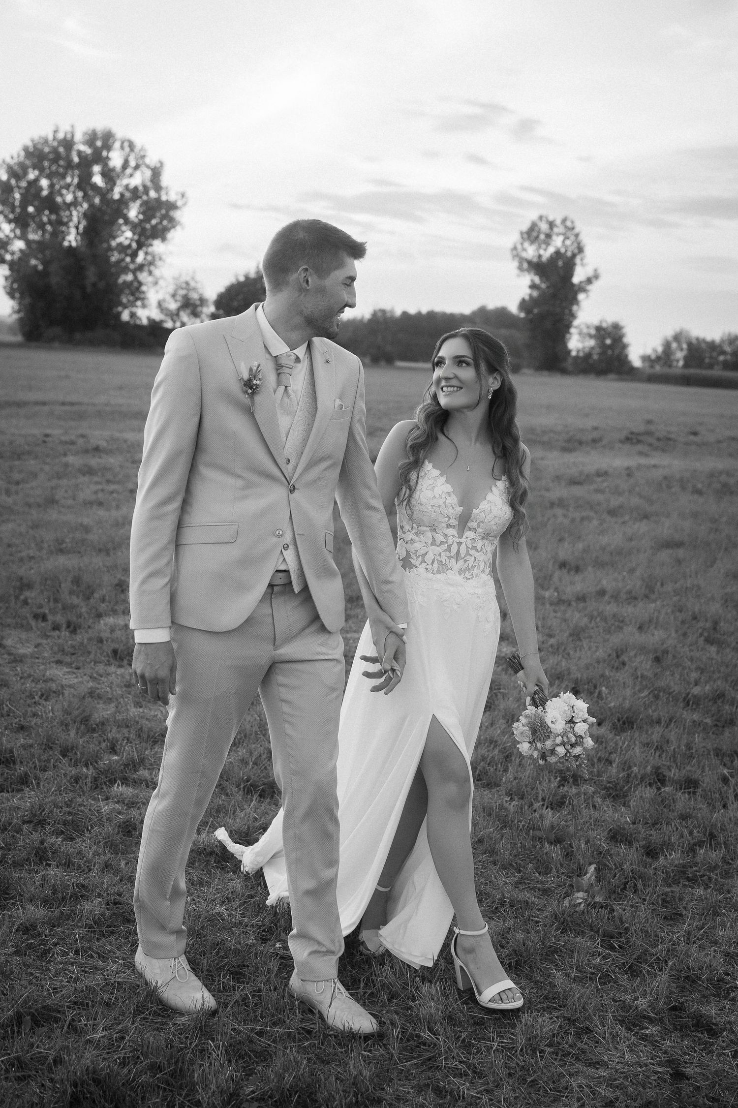 Black and white photo of a bride in a lace gown and groom in a light suit walking hand in hand through a grassy field at sunset.