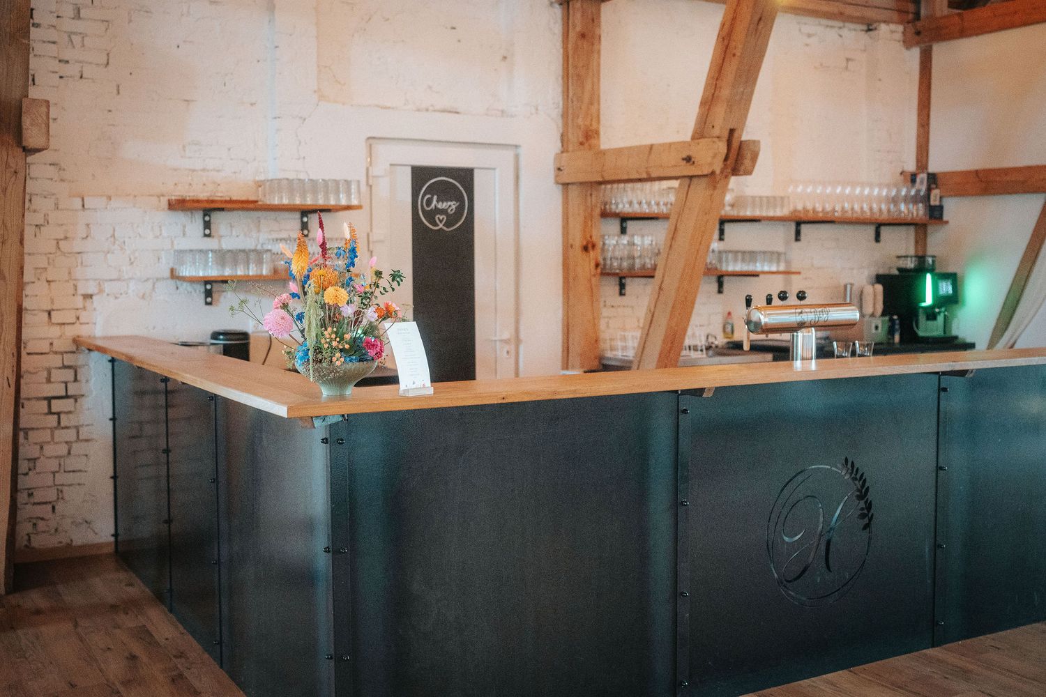 Rustic cafe bar with a dark metal counter, fresh flower bouquet, wooden beams, whitewashed brick walls, and open shelves with glassware.