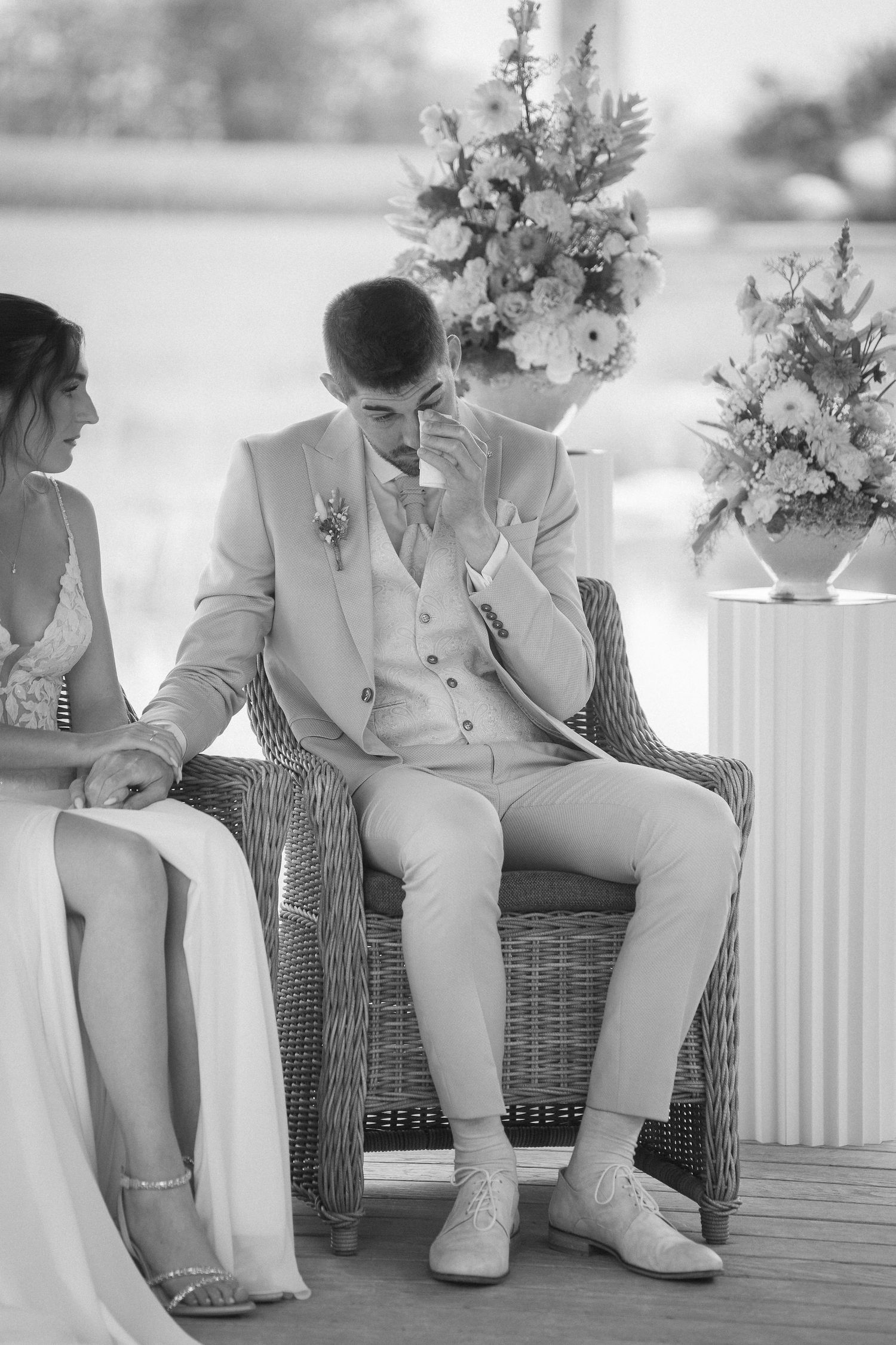 Emotional groom in a light suit wipes away tears while seated in a wicker chair beside his bride at an outdoor wedding ceremony.