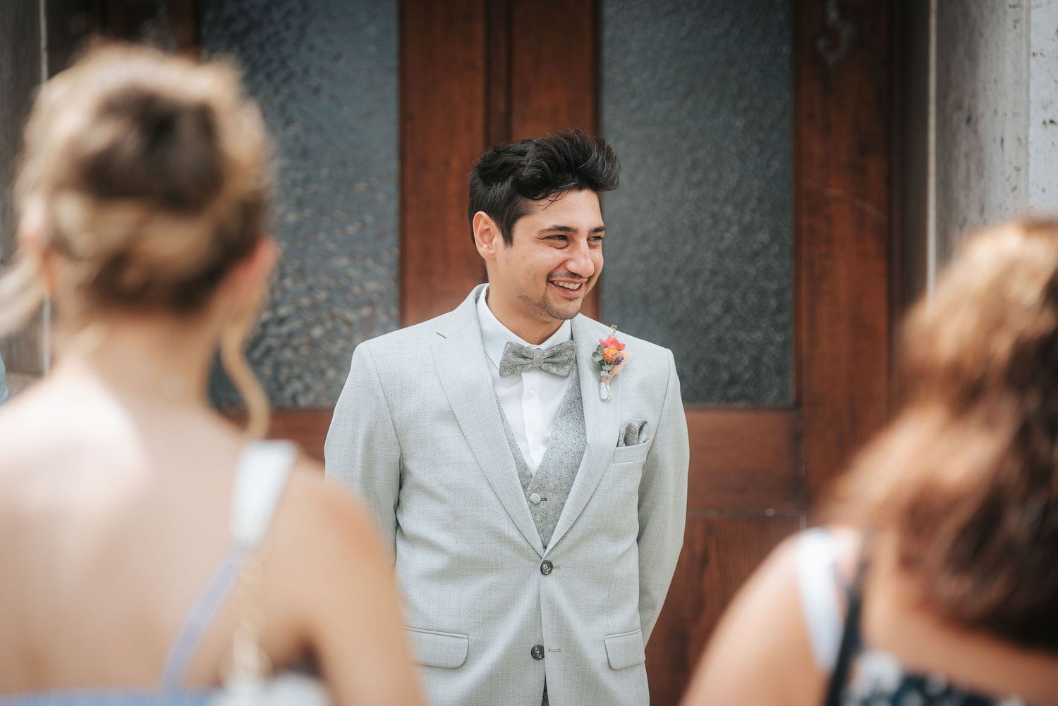 Smiling groom in a light gray three-piece suit with a bow tie and boutonniere stands before a wooden door as two guests face him.