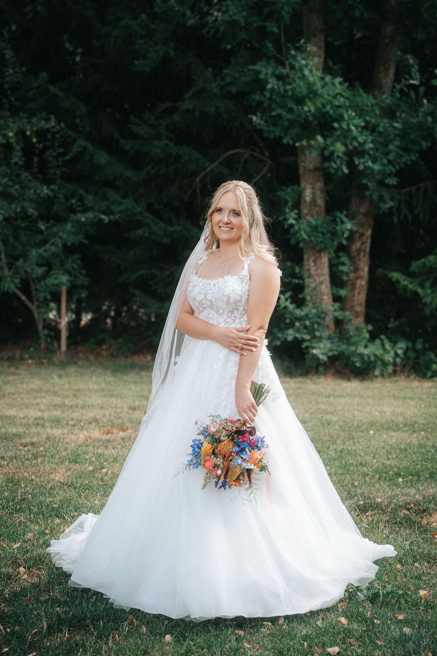 Smiling bride in a white lace-bodice ball gown and veil holding a colorful wildflower bouquet on a sunny lawn with green trees behind.