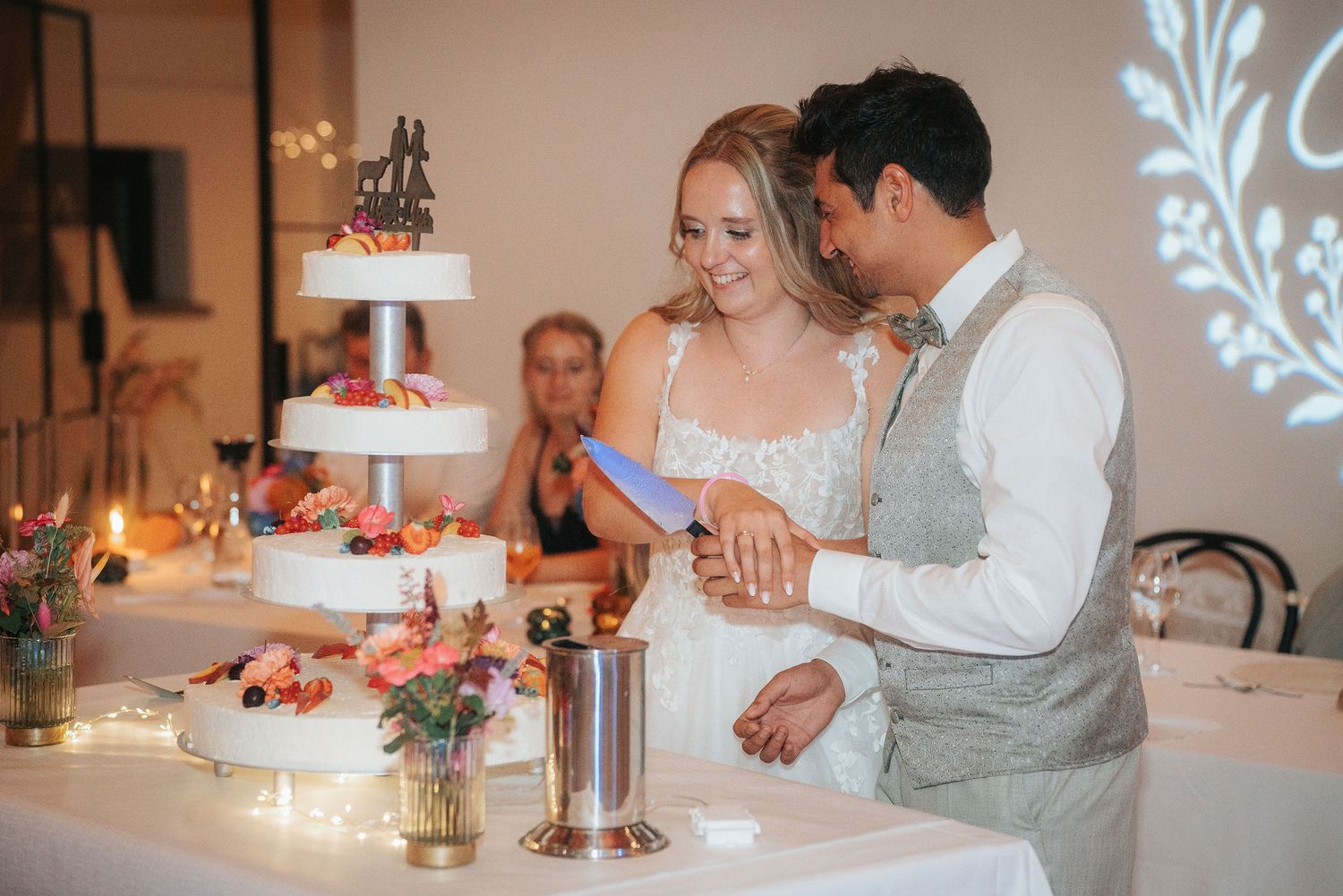 Smiling bride and groom cut their tiered wedding cake together at a candlelit reception decorated with fresh flowers.