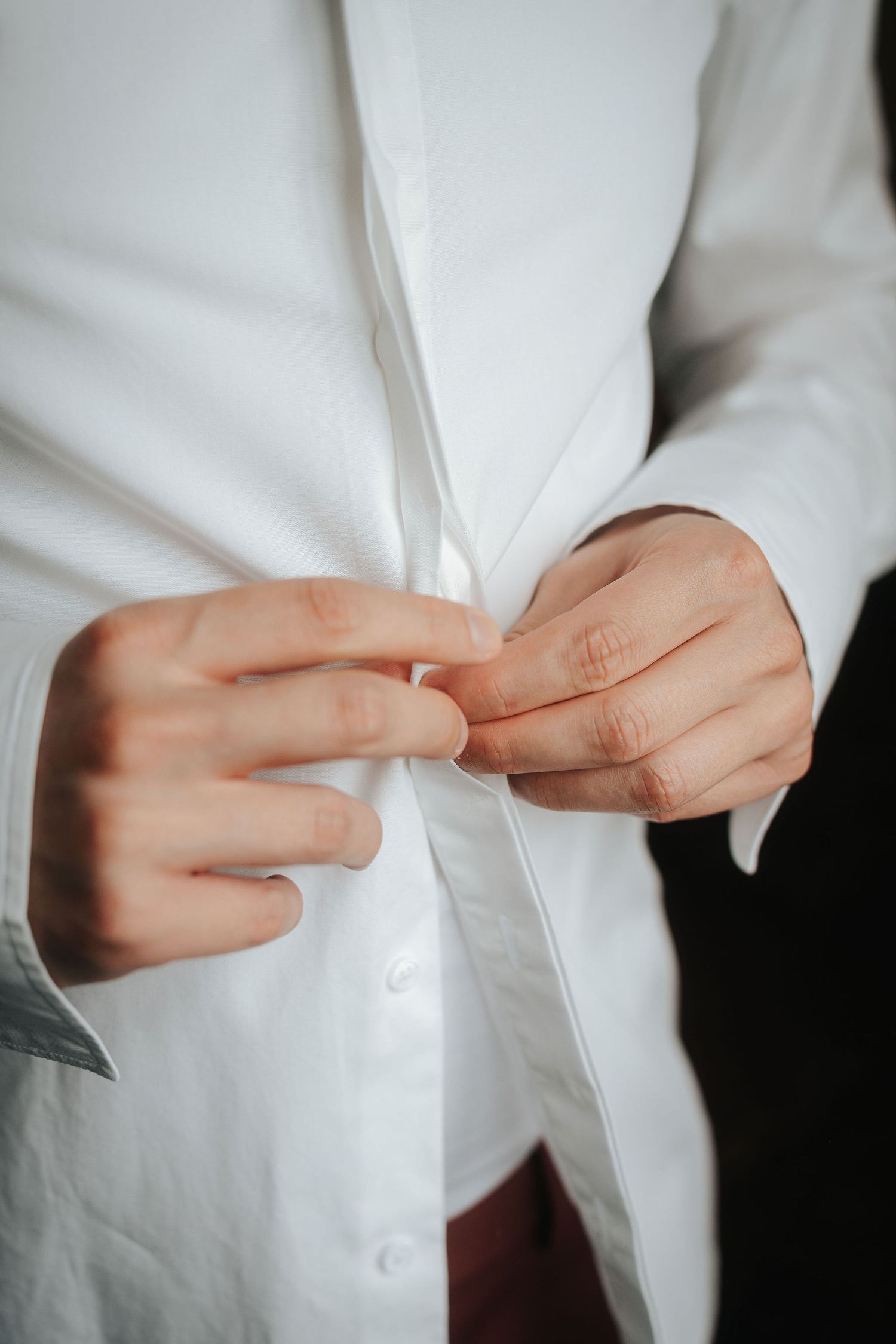 Close-up of a man's hands buttoning a crisp white dress shirt, with dark red trousers visible below, in a getting-ready moment.