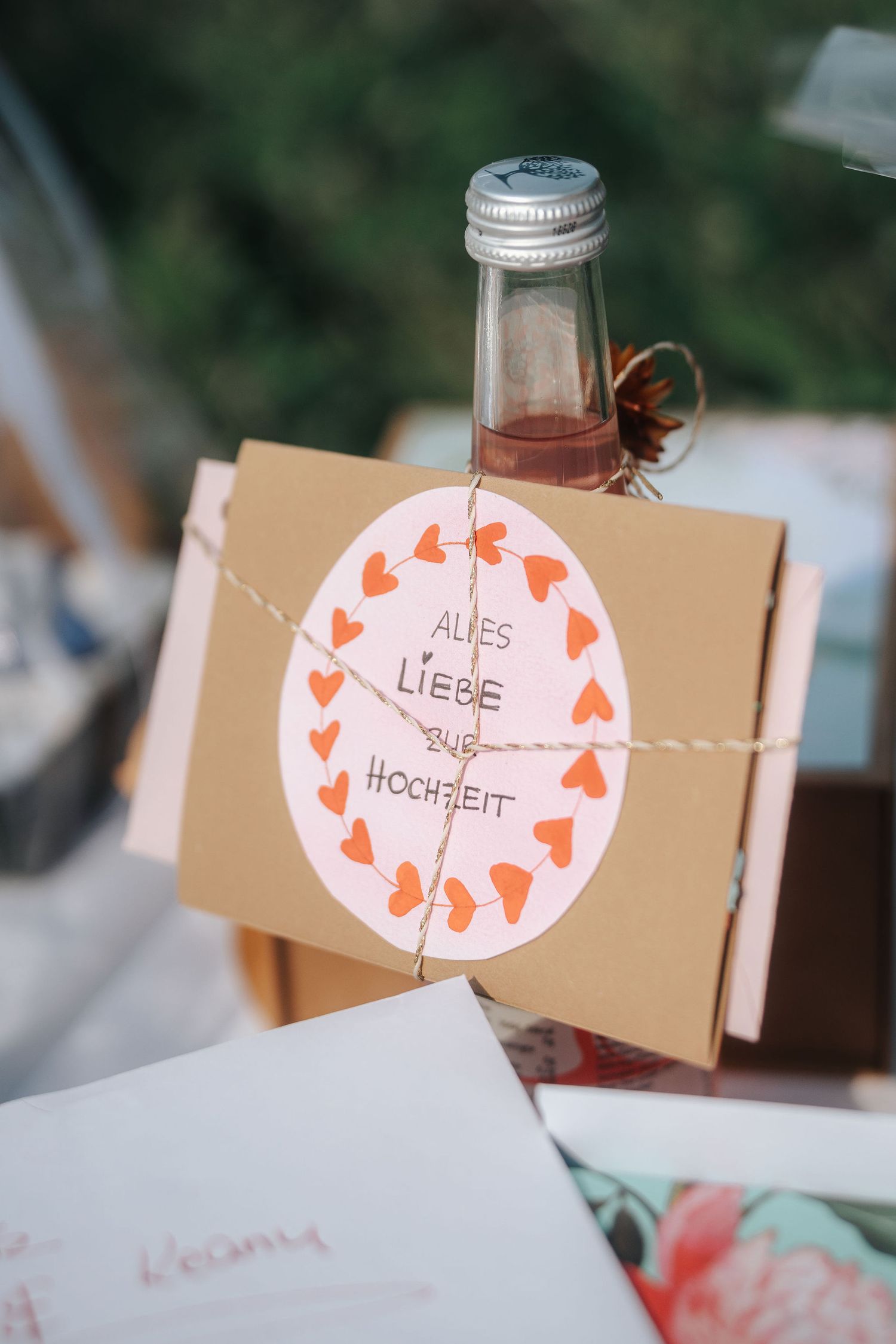 A small bottle tied with twine to a kraft card with orange hearts and German text wishing all the best for the wedding.