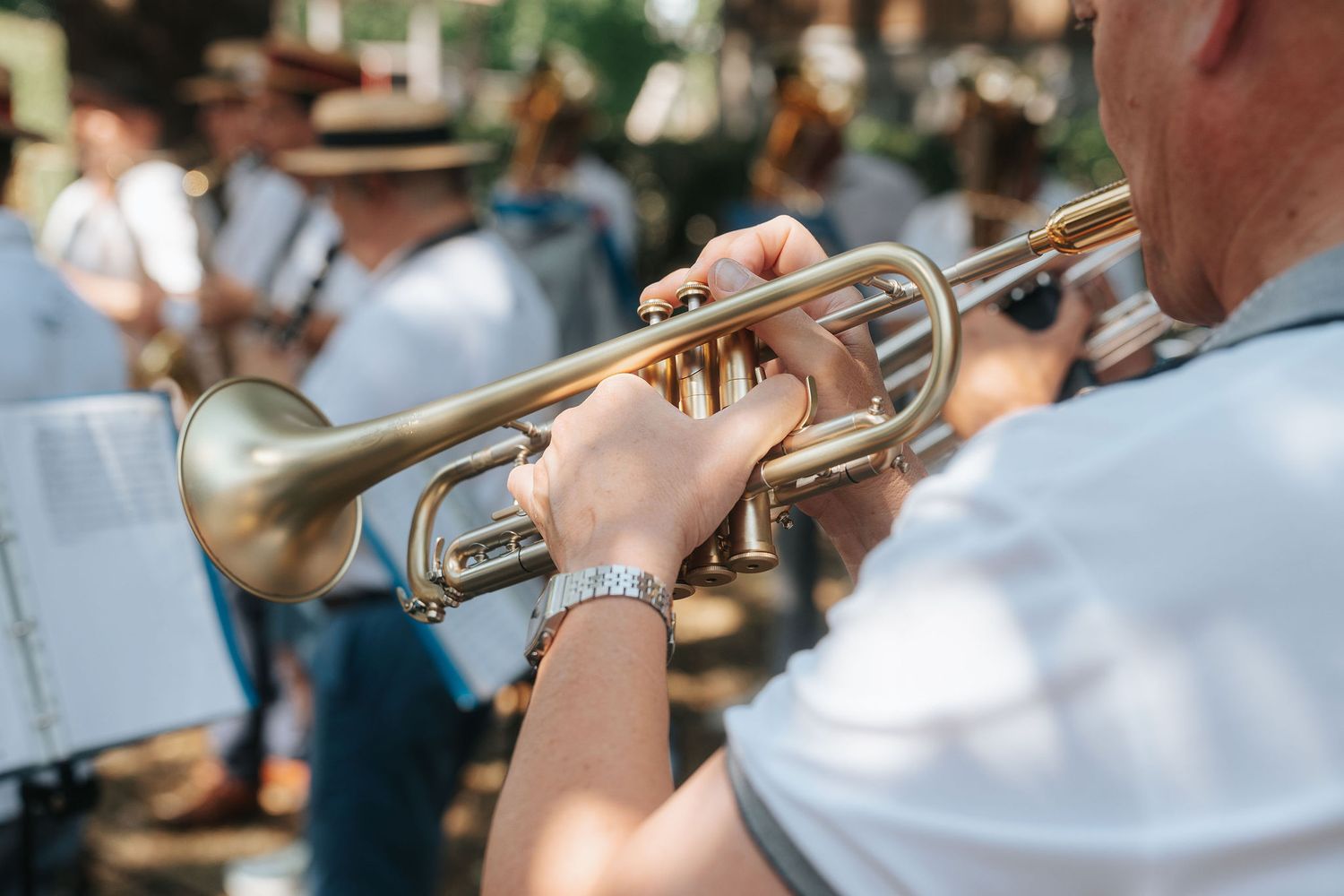 Close-up of a musician in a white shirt playing a brass trumpet outdoors, with a blurred marching band and sheet music in the background.