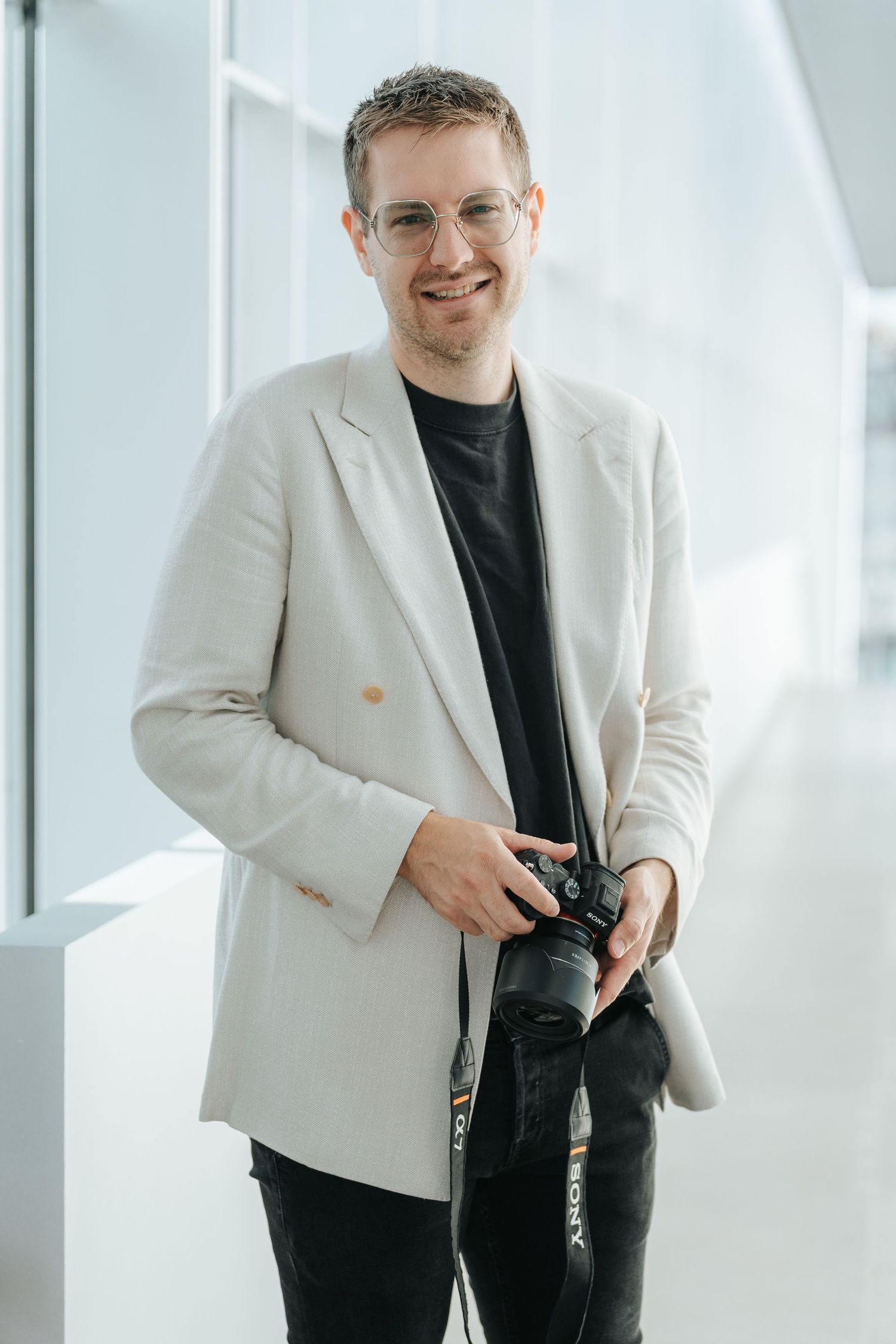 Professional photographer wearing a cream blazer and black t-shirt holds a camera in bright interior setting.