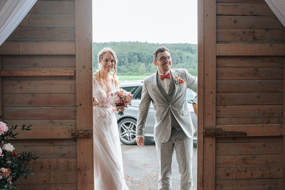 Wedding couple entering rustic barn venue framed by wooden doorway with cars visible in background.