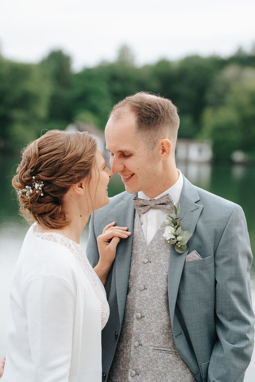 Wedding couple sharing an intimate moment outdoors by a lake, wearing grey suit and white dress with floral details.