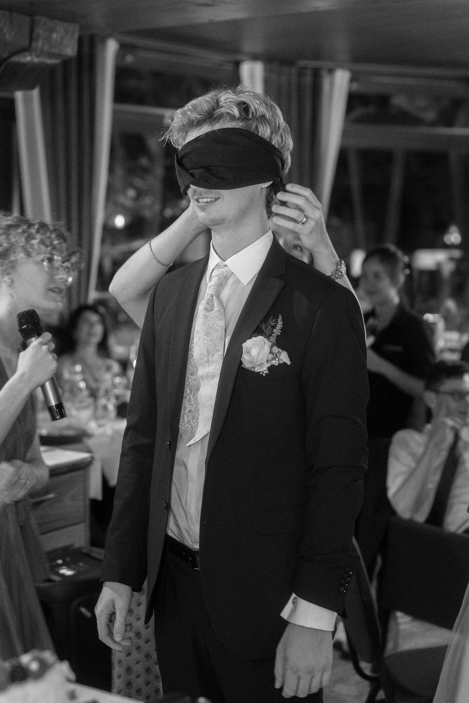 Blindfolded man in a dark suit and boutonniere stands at a wedding reception while a hand covers his eyes and a guest holds a microphone.