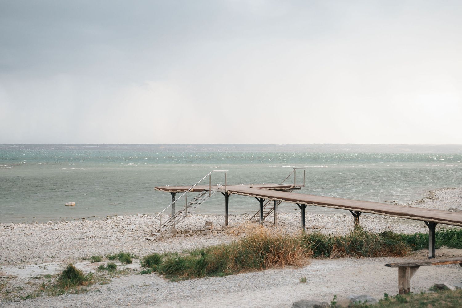 Wooden pier with a ladder extending over a pebbly beach and calm gray sea under an overcast sky, with beach grass in the foreground.
