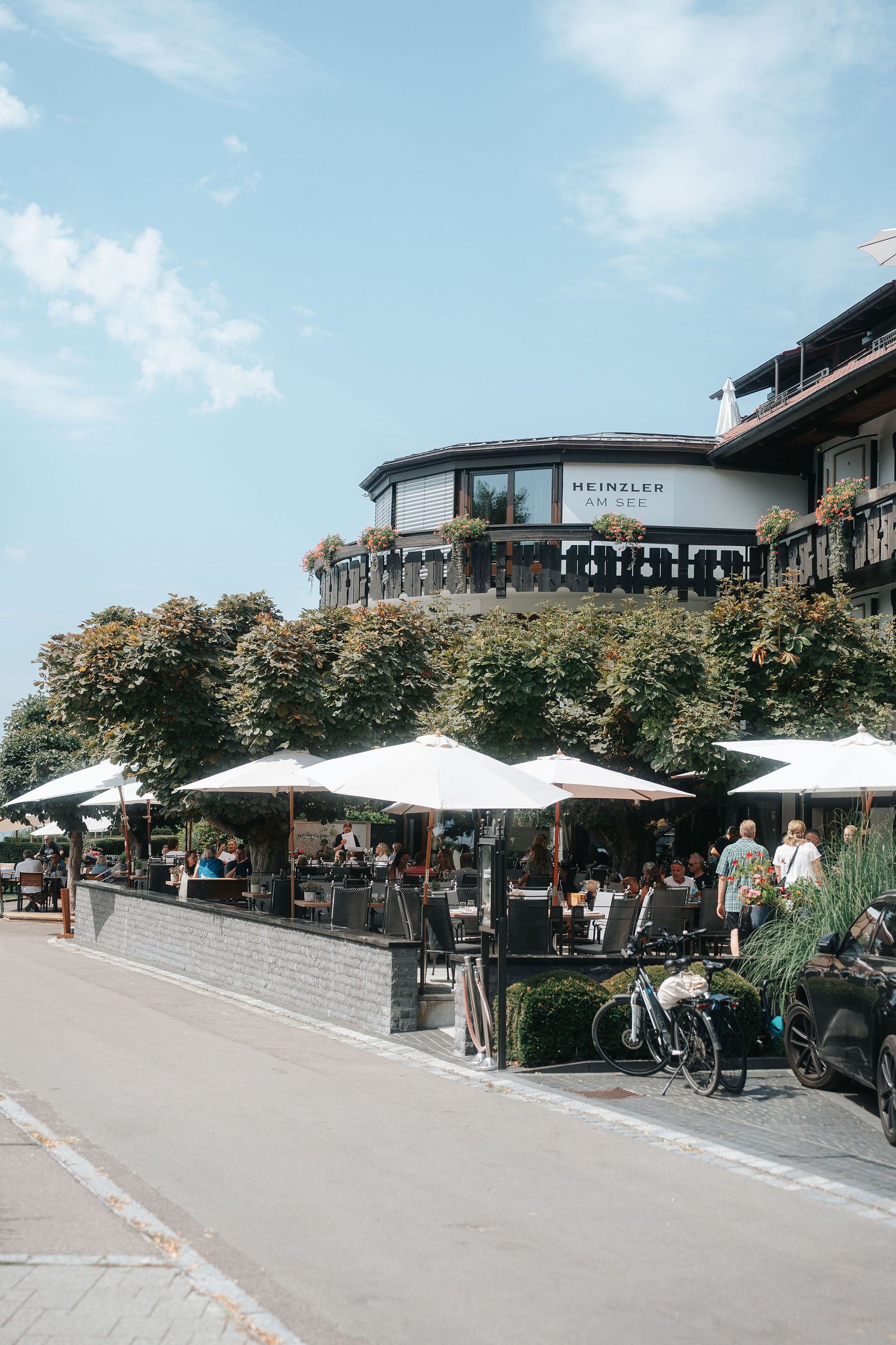 Outdoor restaurant terrace with white parasols and diners beneath leafy trees in front of the Heinzler hotel on a sunny day.