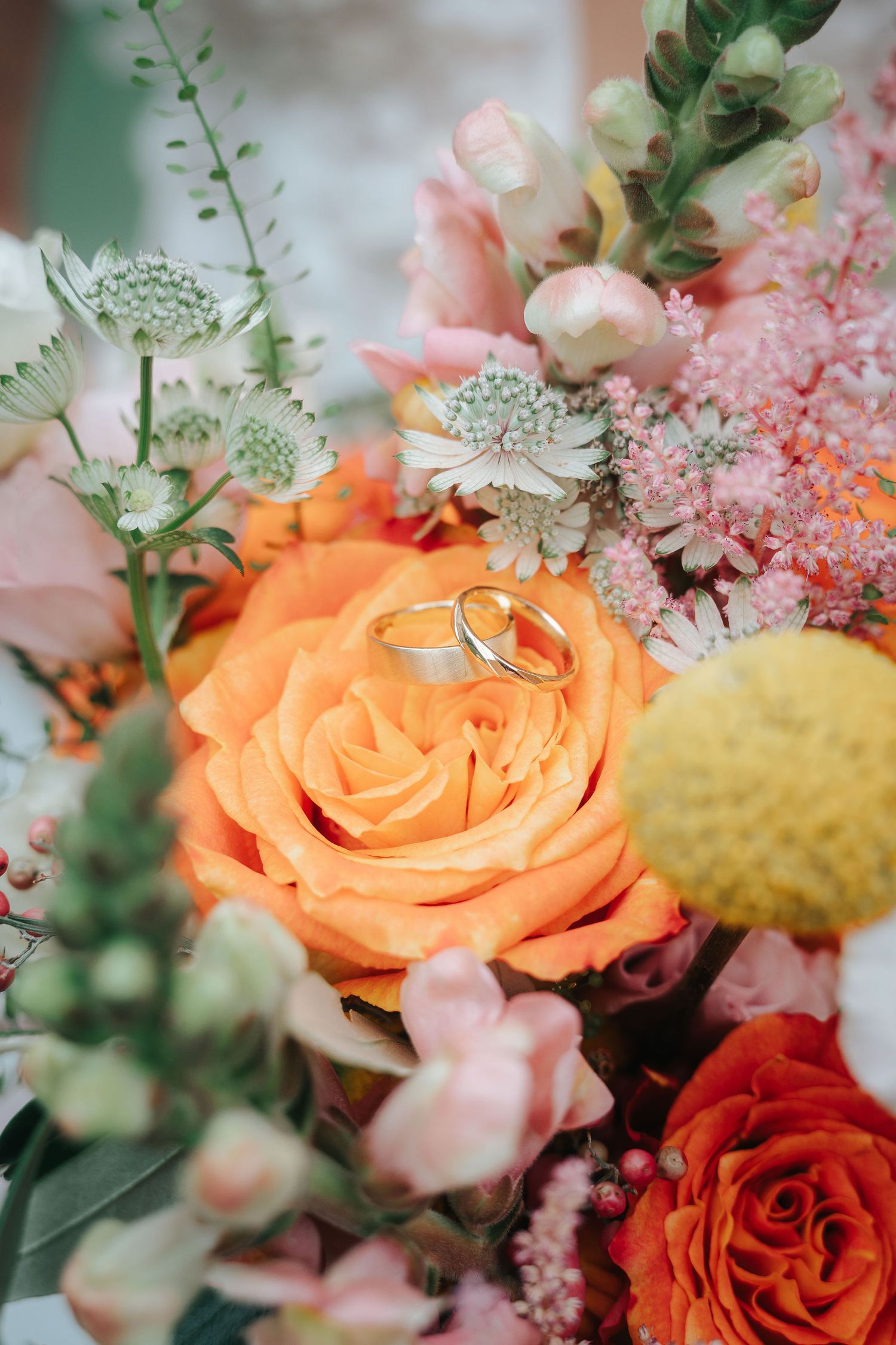 Two wedding rings resting on an orange rose in a colorful bridal bouquet of pink and peach flowers.
