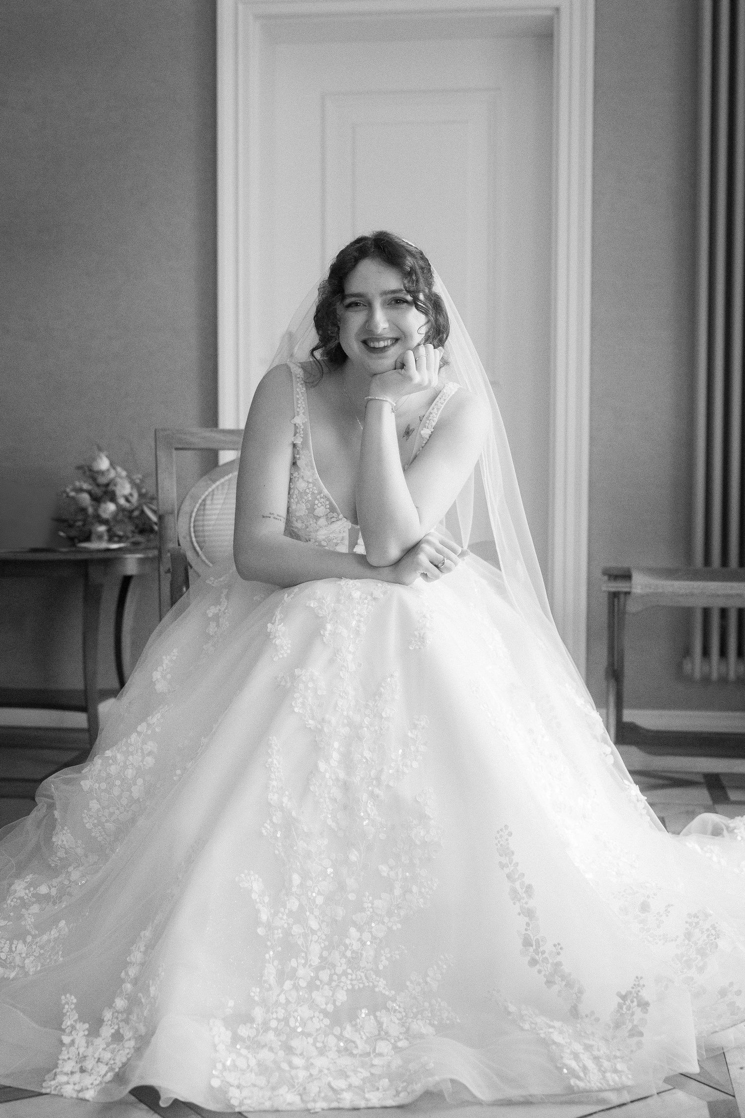 Black and white portrait of a smiling bride in a lace wedding gown and veil, seated in an elegant room with her chin resting on her hand.