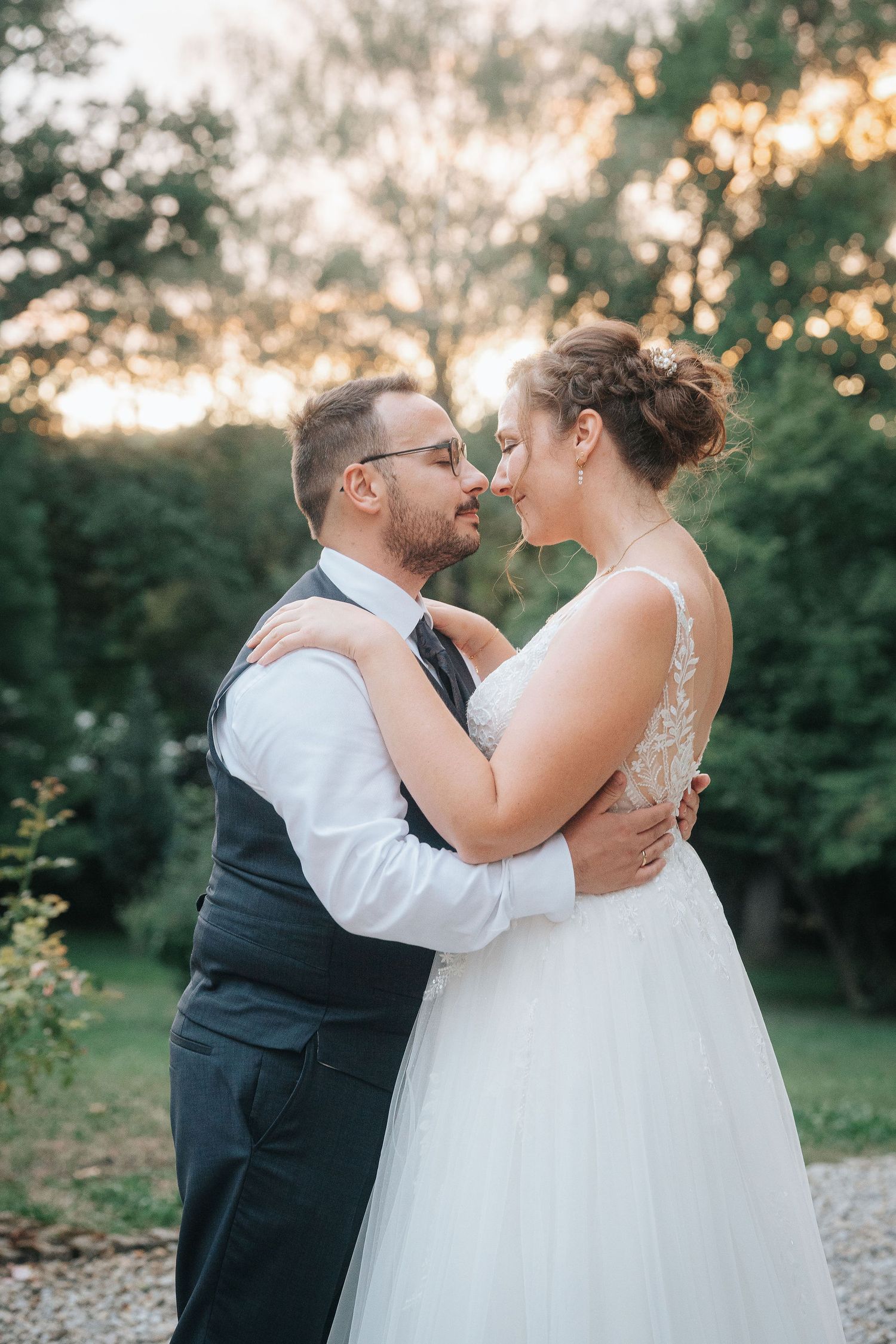 Bride in a lace wedding gown and groom in a vest and tie embrace nose to nose in a sunlit garden at sunset.