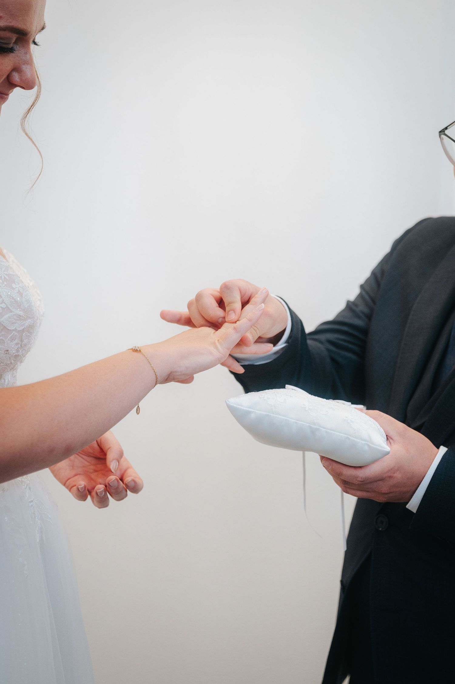 Groom in a dark suit places a wedding ring on the bride's finger during the ceremony, with a white ring pillow in his hand.