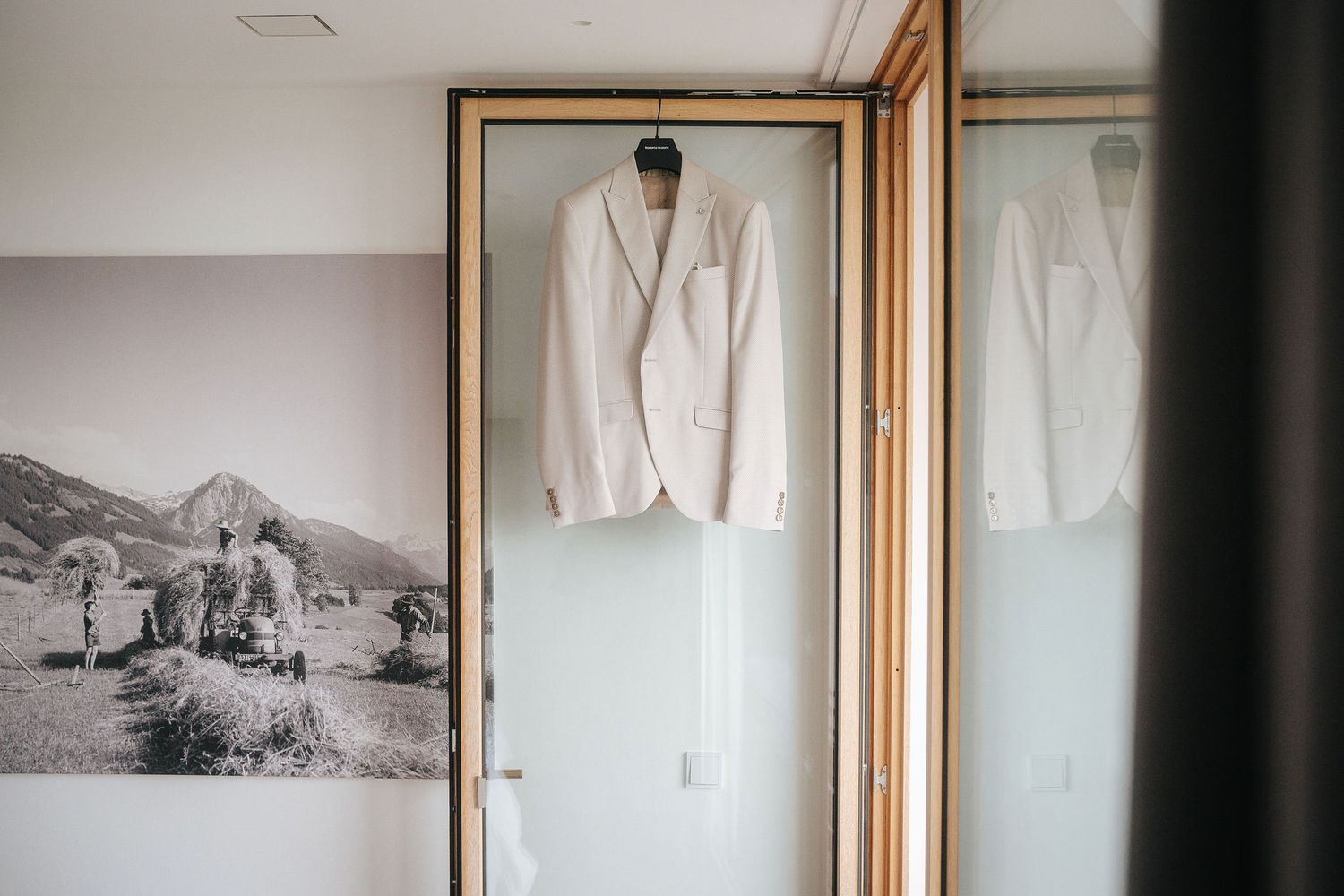 White groom's suit jacket hanging on a wooden-framed glass door beside a black and white mountain landscape photograph.