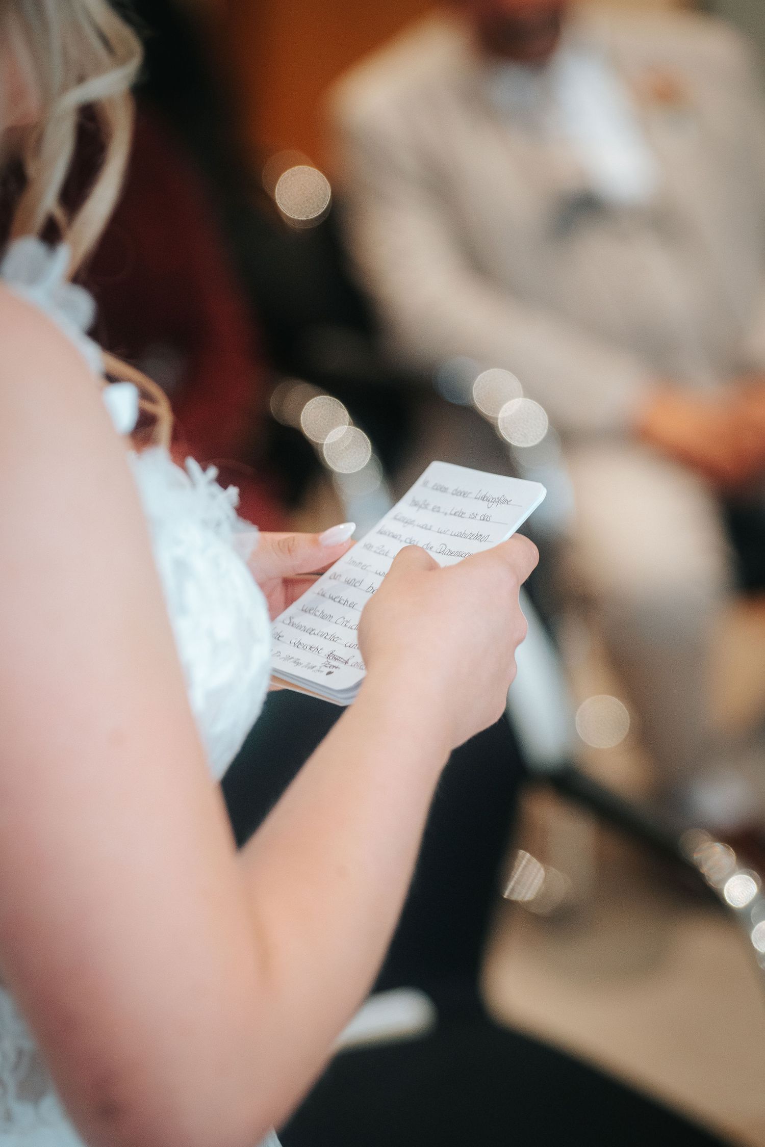 Bride in a white lace dress holds a handwritten note, reading her vows during a wedding ceremony with guests softly blurred behind.