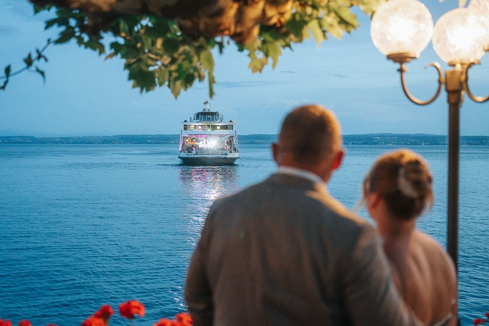 Couple watching illuminated cruise ship sailing on calm blue ocean waters at dusk with glowing lamp post in foreground.