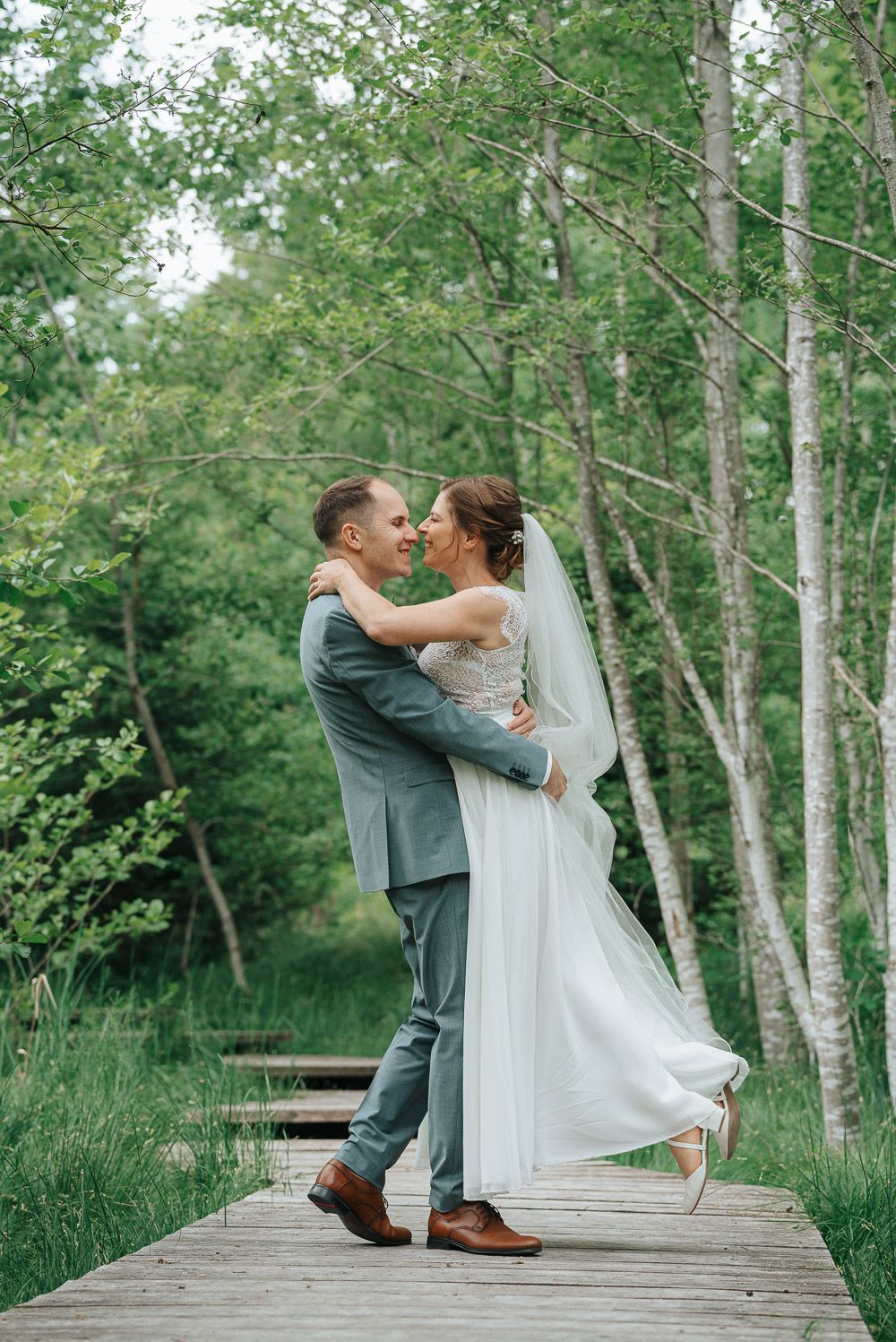 A romantic couple on their wedding day share a tender moment together on a wooden boardwalk surrounded by birch trees.