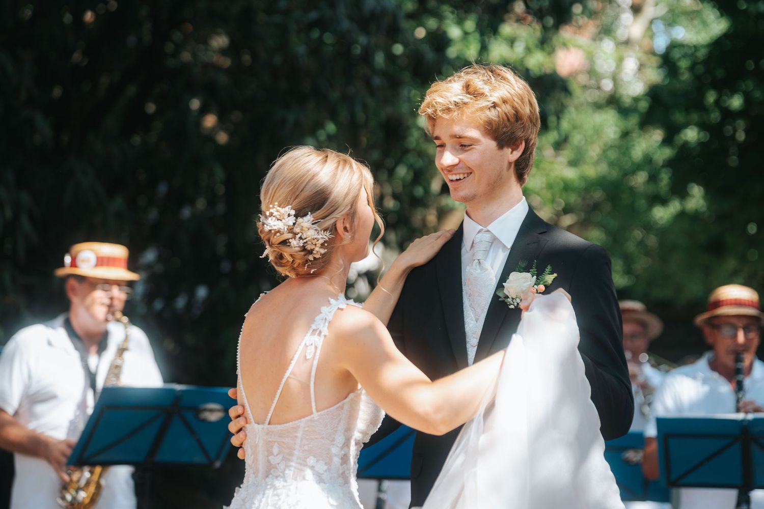 A bride in a lace gown with a floral hair accessory dances with a smiling groom in a dark suit as a jazz band plays outdoors.